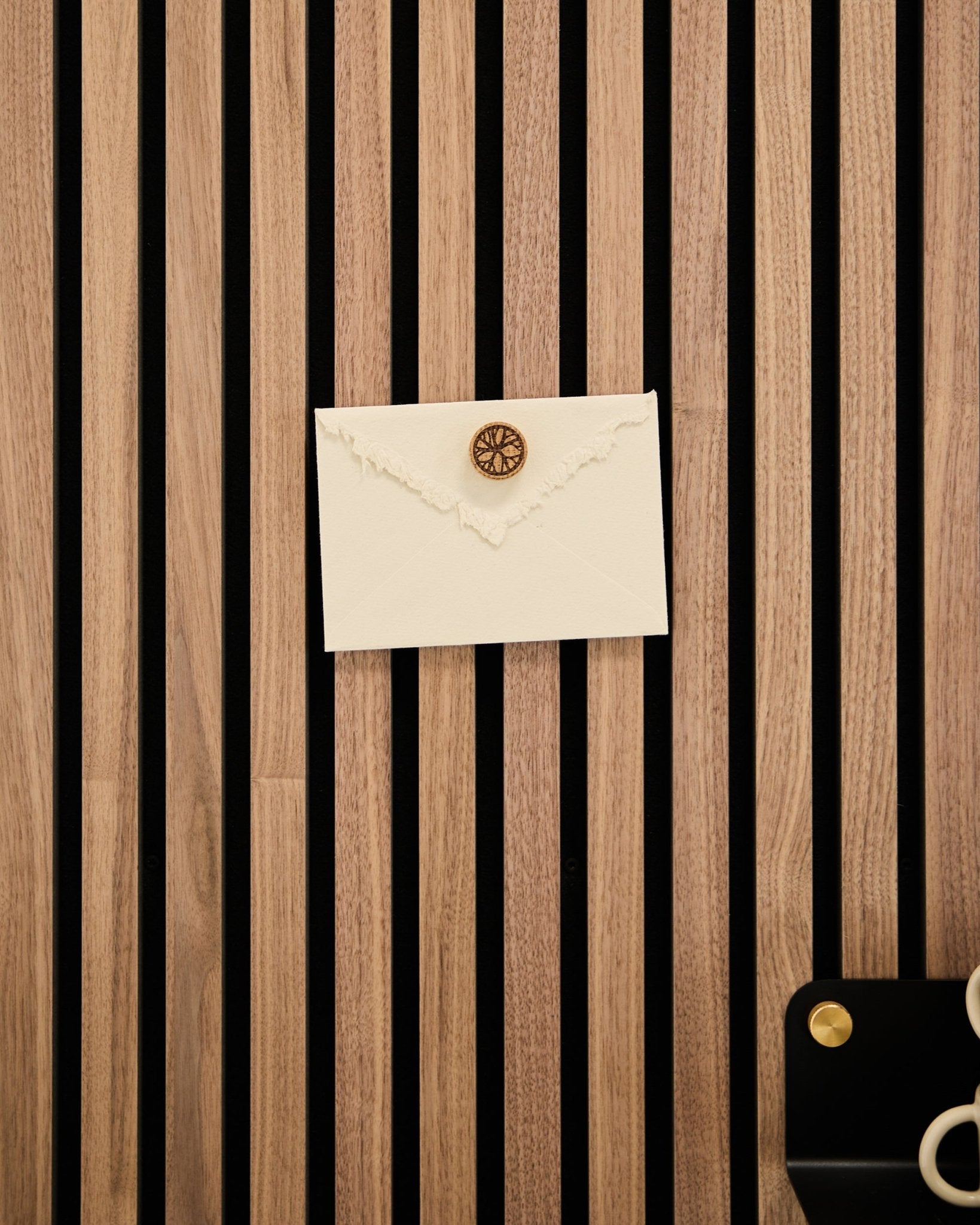 Um envelope creme com selo de cera está fixado a um painel ripado acústico em carvalho da WoodUpp com o conjunto Poster Magnet Norr em Natural Oak; tesoura e tabuleiro nas proximidades.