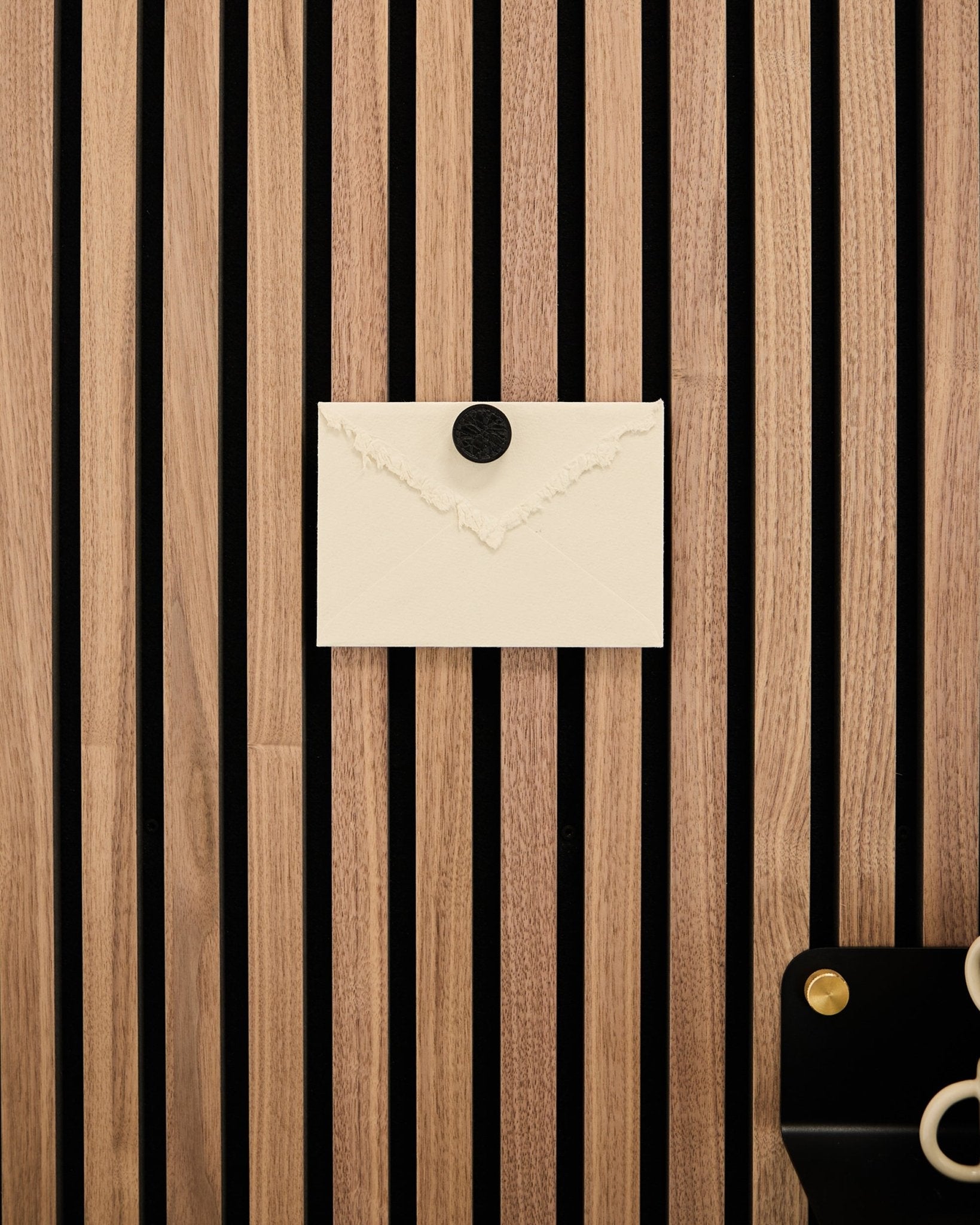 Um envelope creme está preso a um painel acústico de ripas de carvalho Akupanel da WoodUpp Painel acústico com o conjunto de ímanes para posters Norr em Black Oak; há uma tesoura no canto inferior direito.