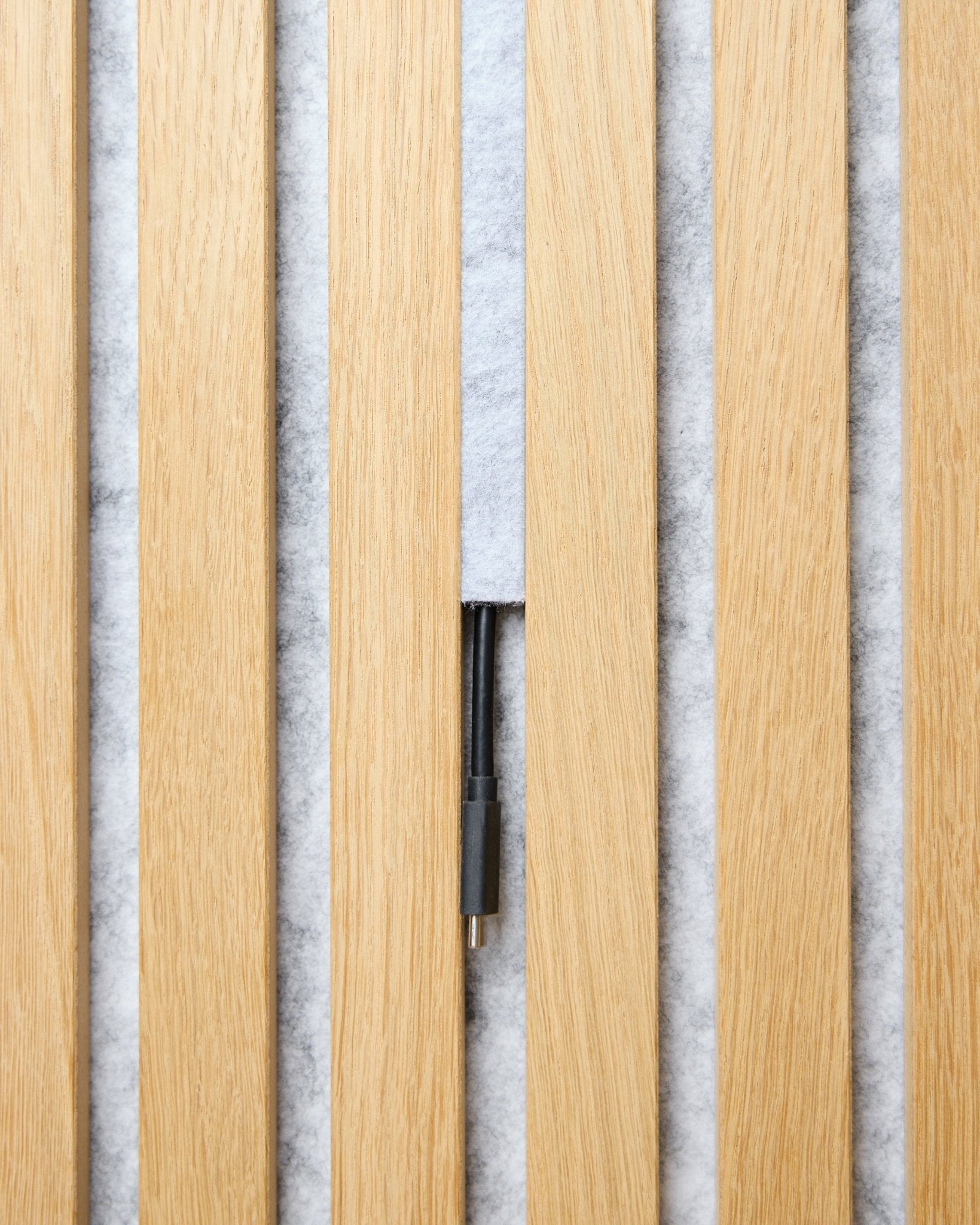 O conjunto Dupps Pop-on Cable Cover (grey, 3 unid.) da WoodUpp organiza cabos entre ripas acústicas de madeira clara Akupanel, sobre um fundo branco texturado.