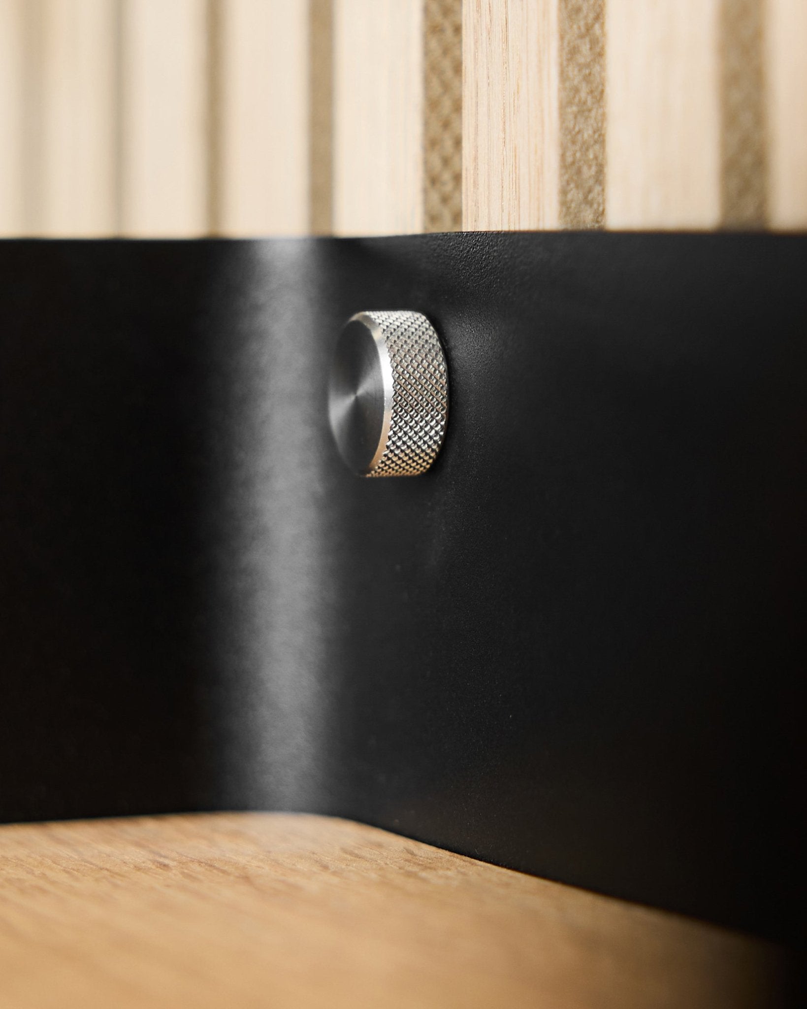 Grande plano de dois parafusos de aperto manual WoodUpp (M5x10 mm, 20x8.8 em aço inoxidável steel) sobre uma superfície metálica black, com ripas acústicas de madeira Akupanel em segundo plano.