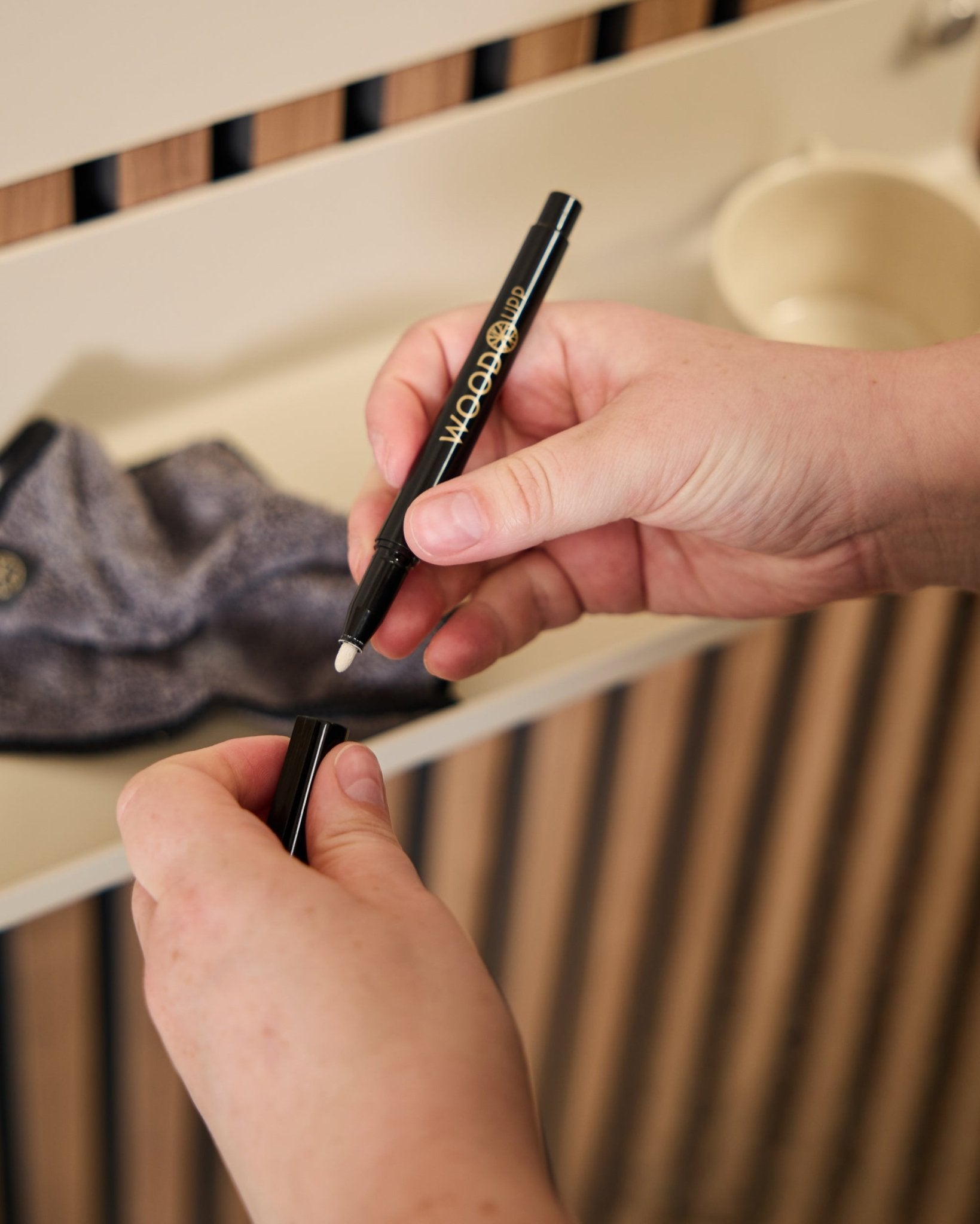 Uma pessoa segura um marcador Norr | Blackboard set aberto da WoodUpp ao lado de um pano de microfibra e de uma chávena beige, produtos pensados para usar com painéis acústicos de madeira.