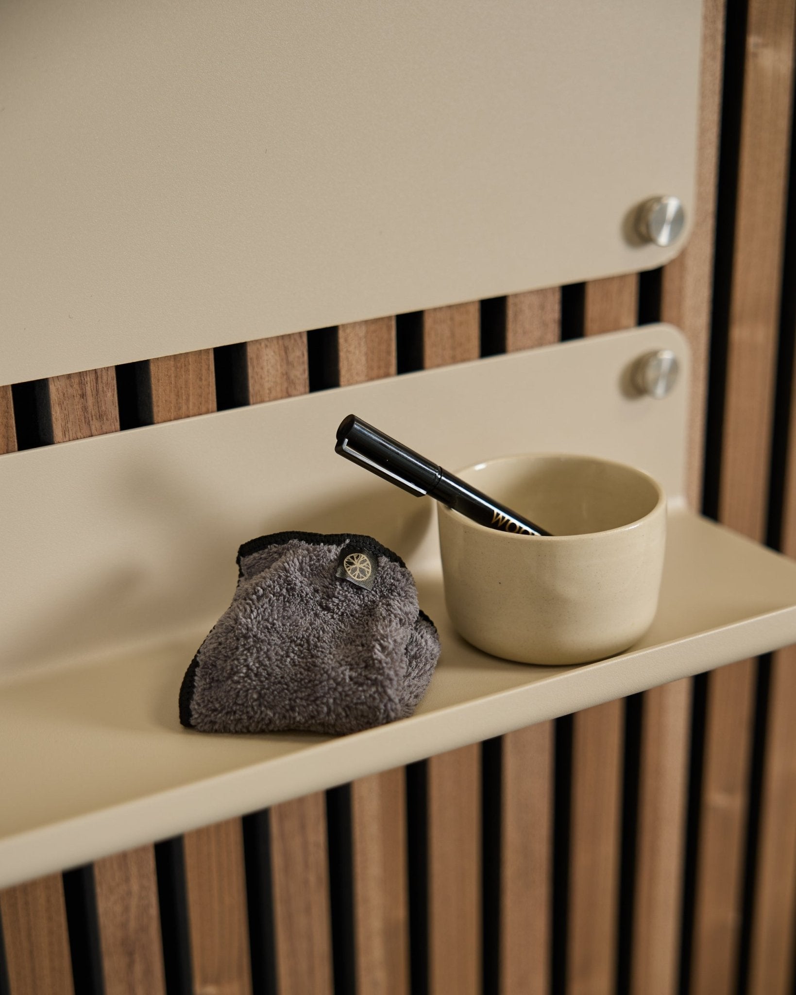 O conjunto Blackboard Norr da WoodUpp inclui uma chávena small beige, uma escova com cabo black e um pano de limpeza sobre uma prateleira beige com painel acústico de madeira em estilo nórdico.