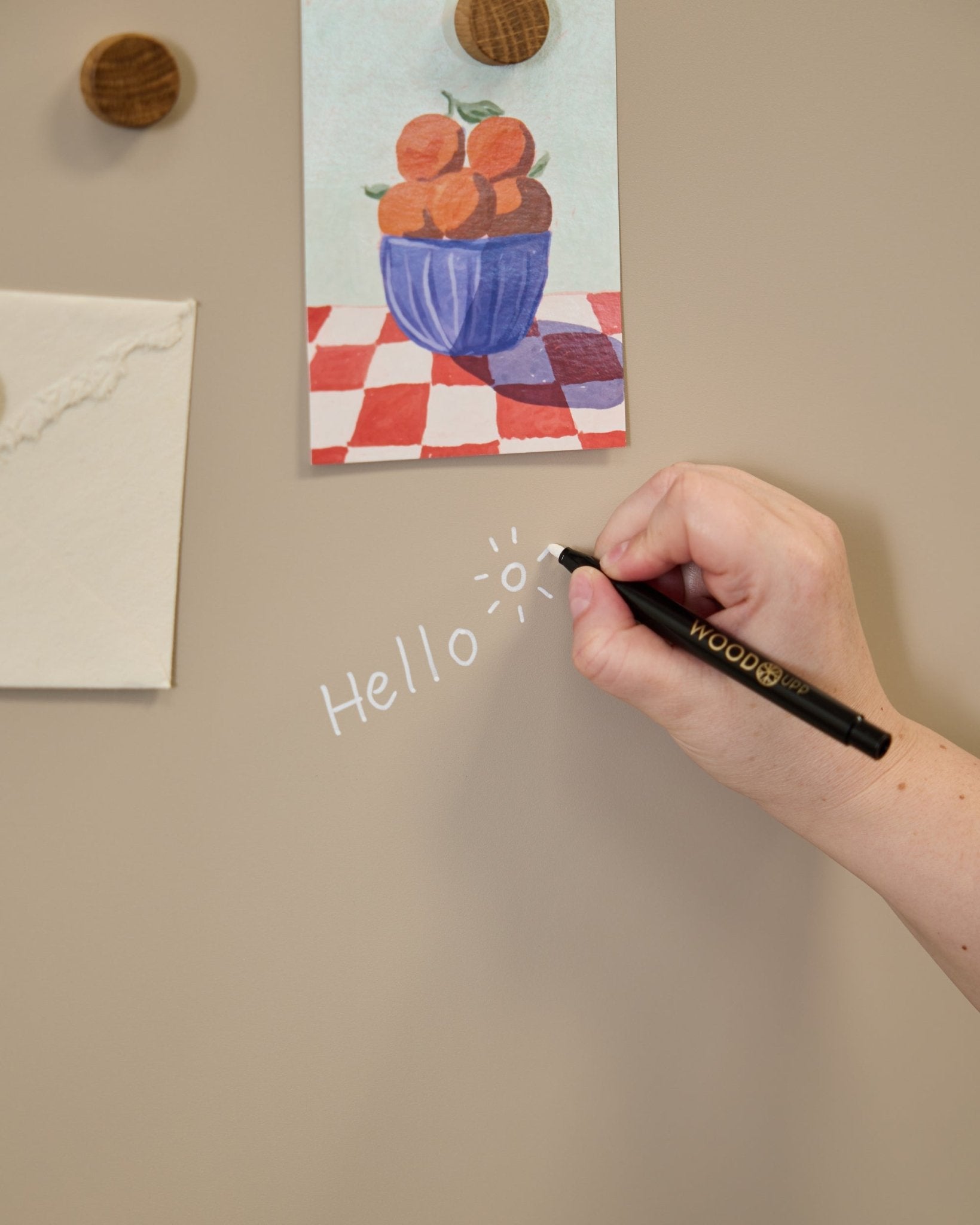 Uma mão escreve hello com um desenho de sol no Blackboard Norr da WoodUpp, colocado ao lado de arte presa com ímanes redondos num(a) Painel acústico.