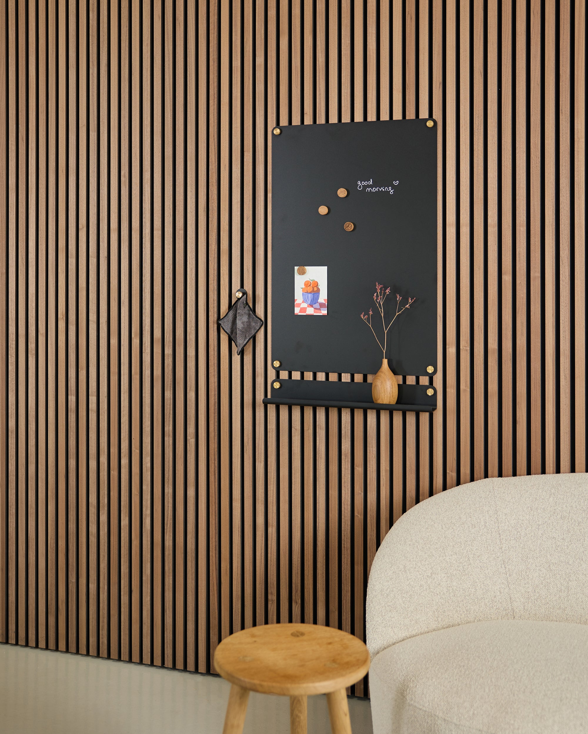 A room with a beige armchair, wooden stool, and a black wall board with magnets and photos displayed on a wood Akupanel acoustic slatwall panel.
