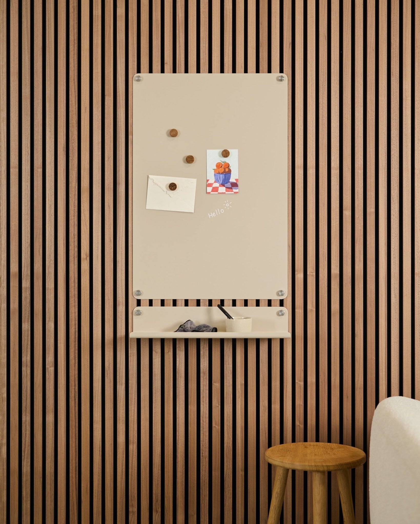 Quadro Magnético Beige Norr da WoodUpp montado por cima de uma prateleira small numa parede com painel acústico de madeira Akupanel, com um banco e um sofá branco por perto.