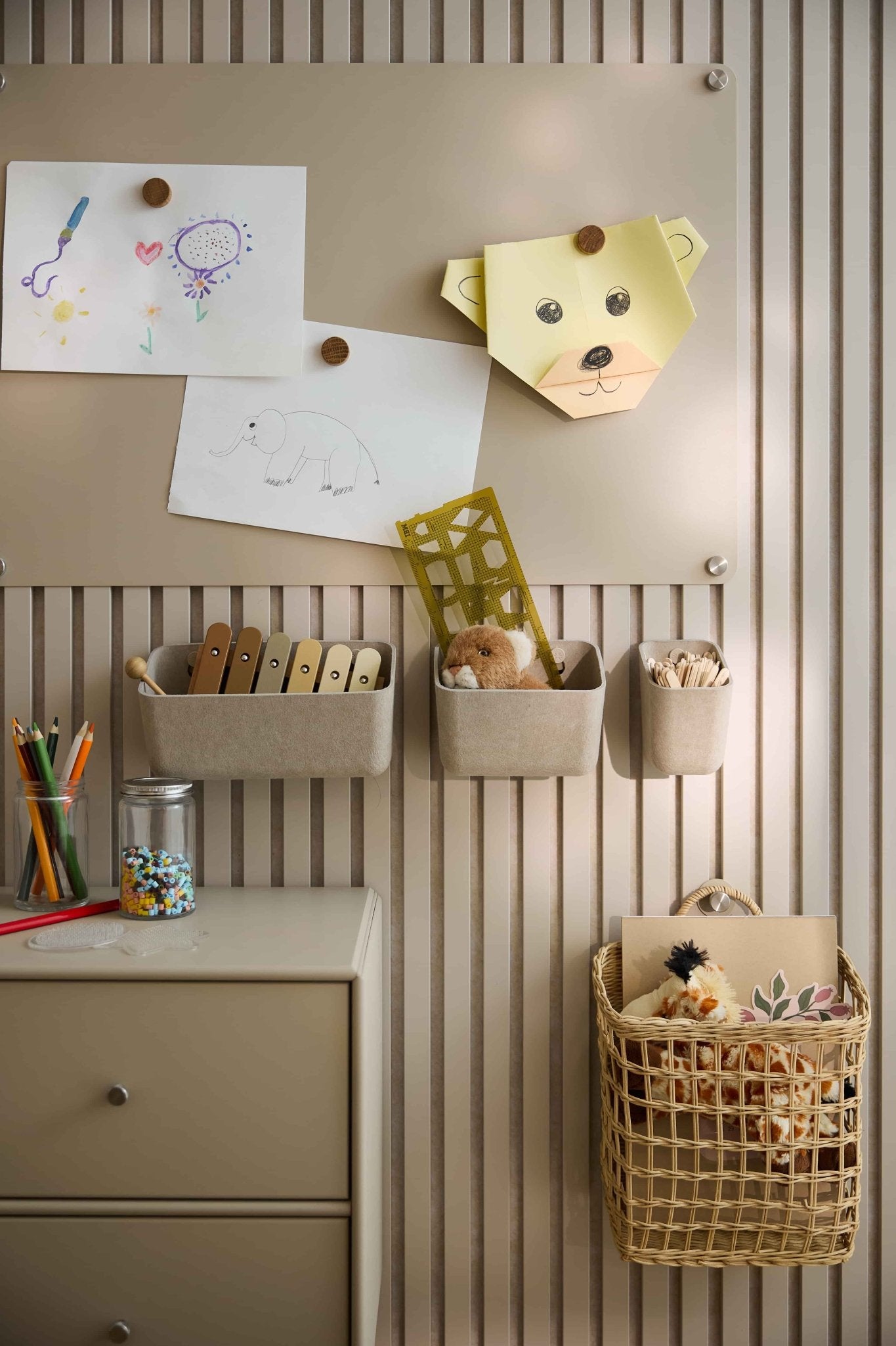 As obras de arte e os brinquedos das crianças estão dispostos numa parede bege com um painel acústico WoodUpp, que inclui recipientes de feltro, cestos, uma gaveta e um painel perfurado.