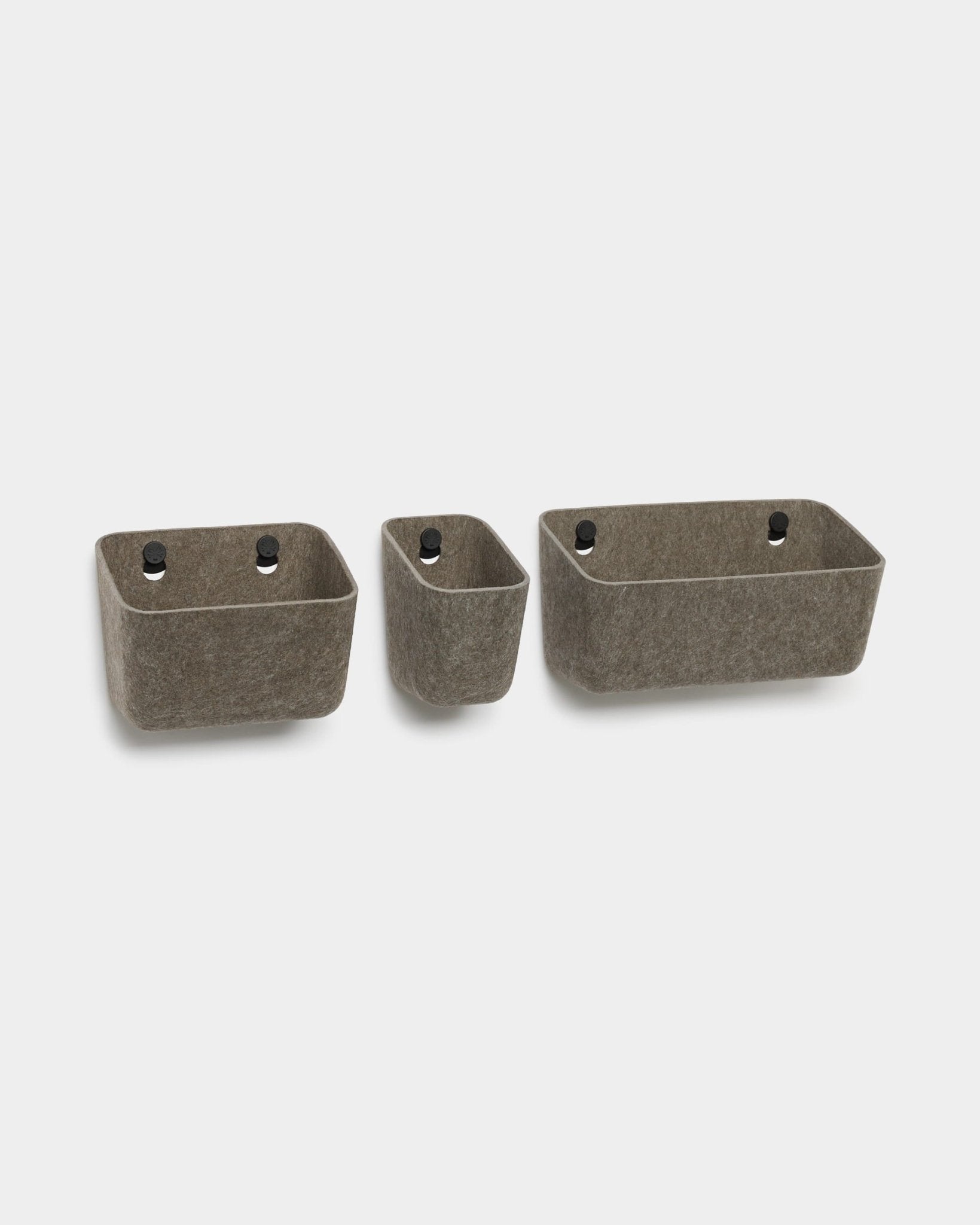 Três recipientes de feltro WoodUpp Luva em cinzento escuro quente com ilhós pretos, concebidos para montagem na parede como organizadores compatíveis com os painéis acústicos WoodUpp.