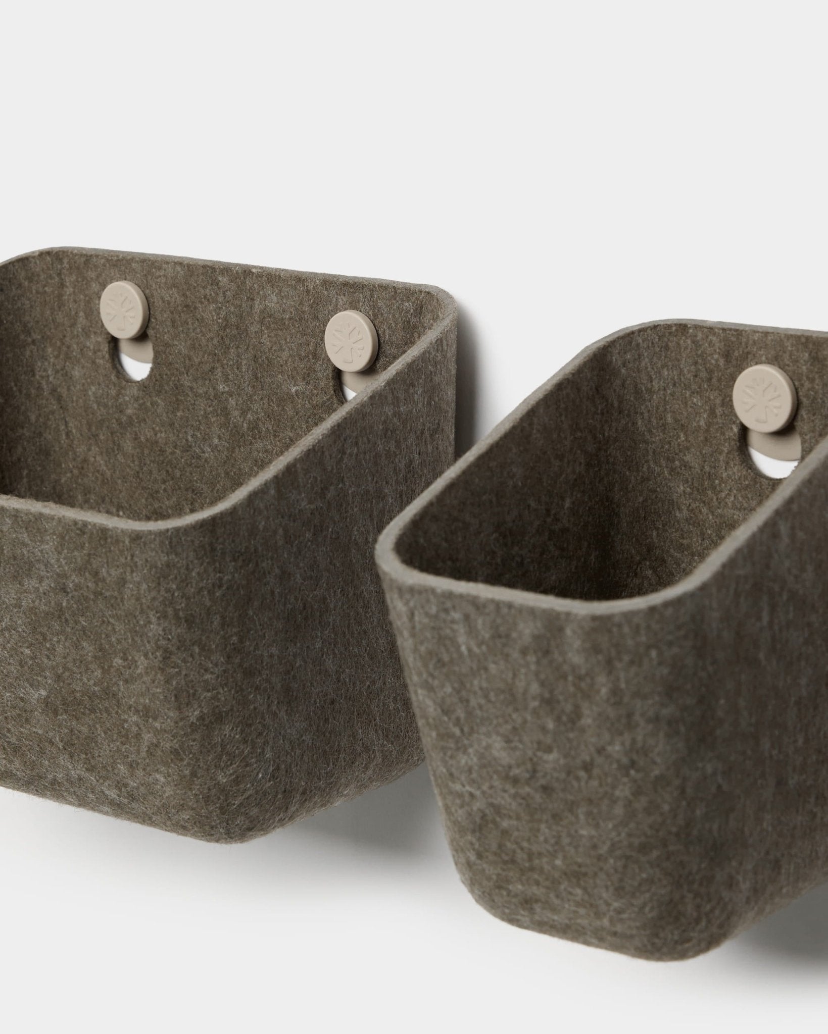 Dois recipientes de feltro Luva, em cinzento escuro e quente, da marca WoodUpp, estão pendurados em ganchos bege fixados a um painel acústico, proporcionando um espaço de arrumação minimalista na parede com cantos arredondados.