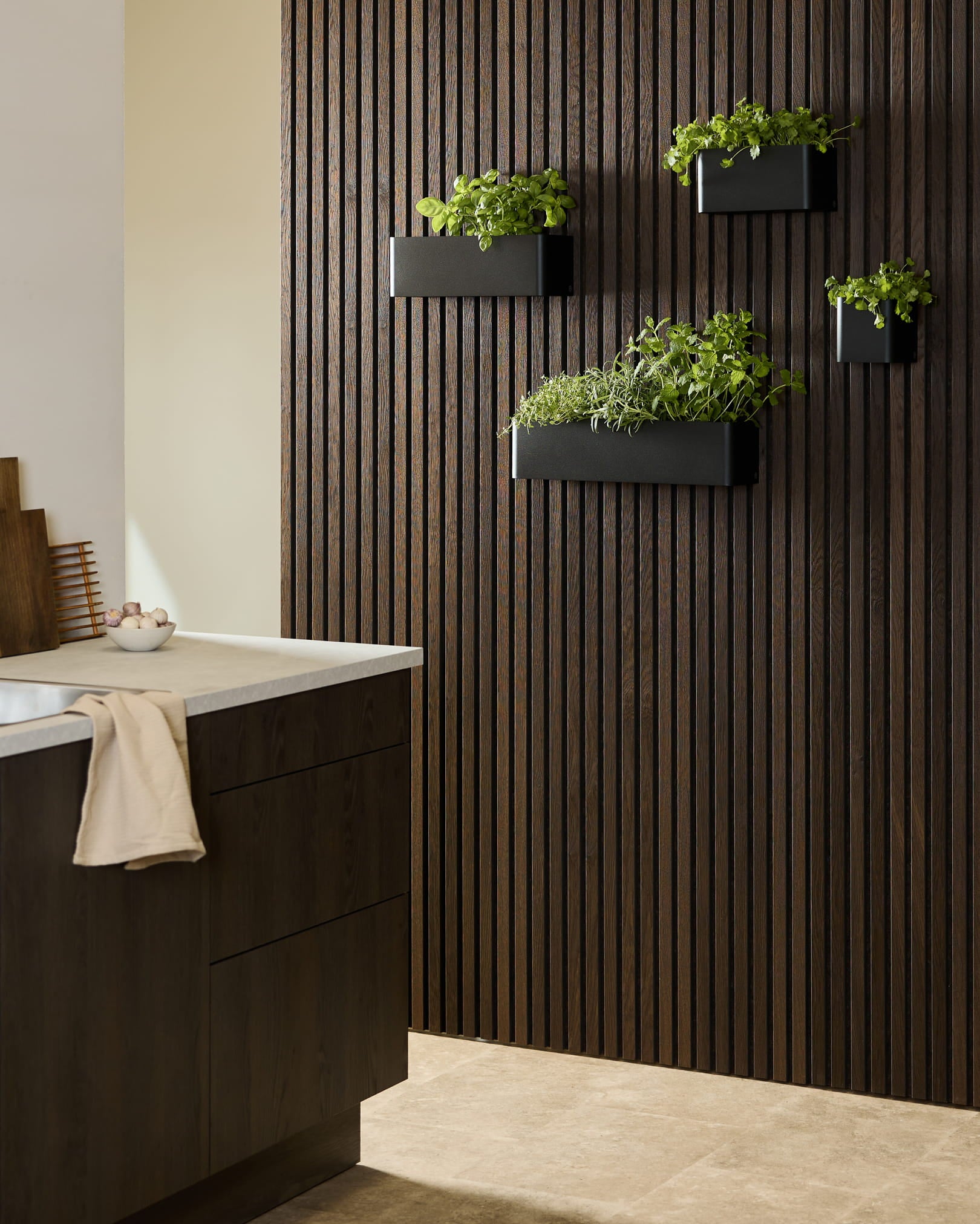 Cozinha moderna com um painel de madeira escura WoodUpp Akupanel Painel acústico, Plant Box Norr black com ervas aromáticas e armários escuros com tampo branco.