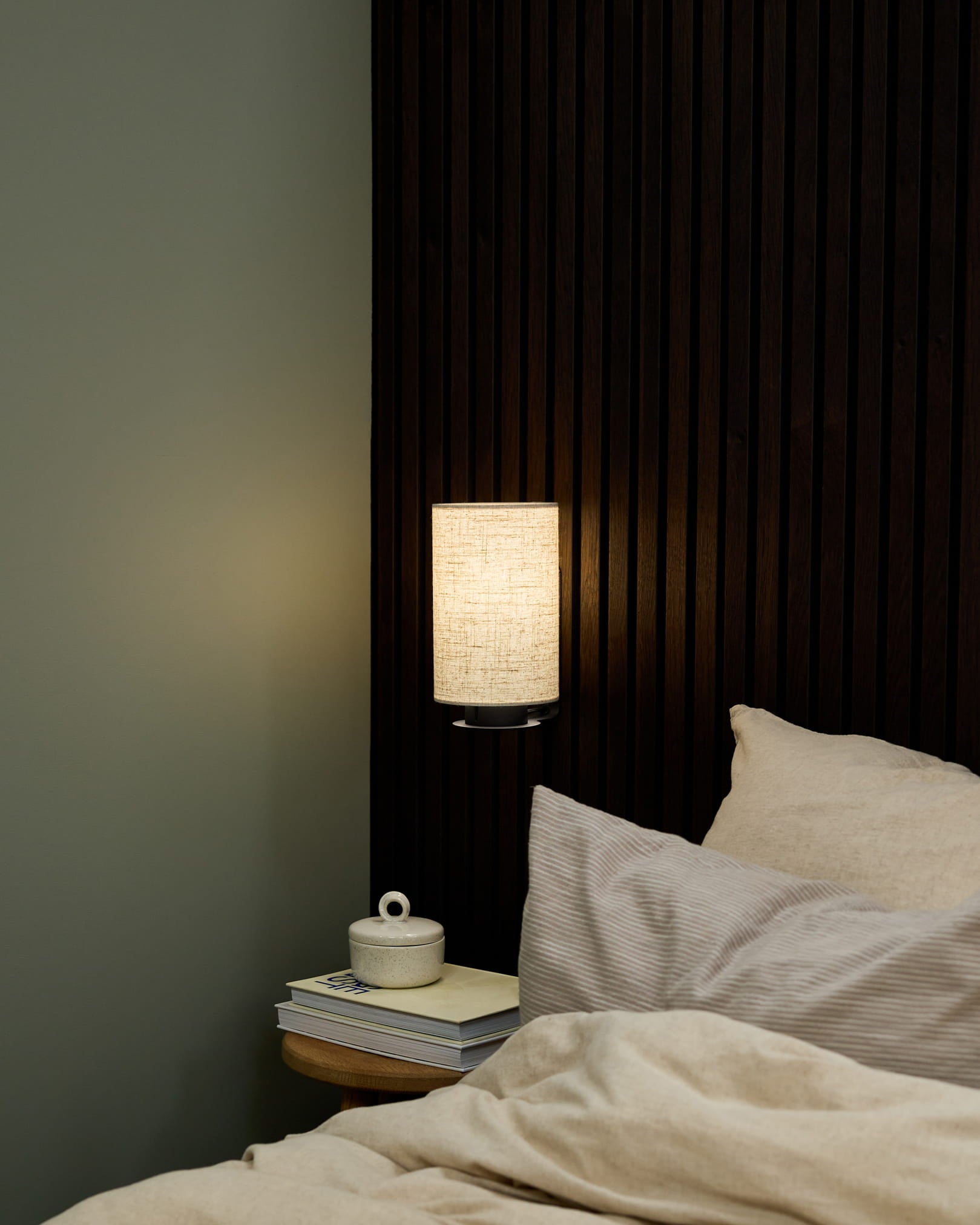Canto de quarto com um candeeiro de parede Feya da WoodUpp em Black/Beige, roupa de cama em beige e uma mesa de cabeceira com livros e uma chávena de cerâmica com tampa.
