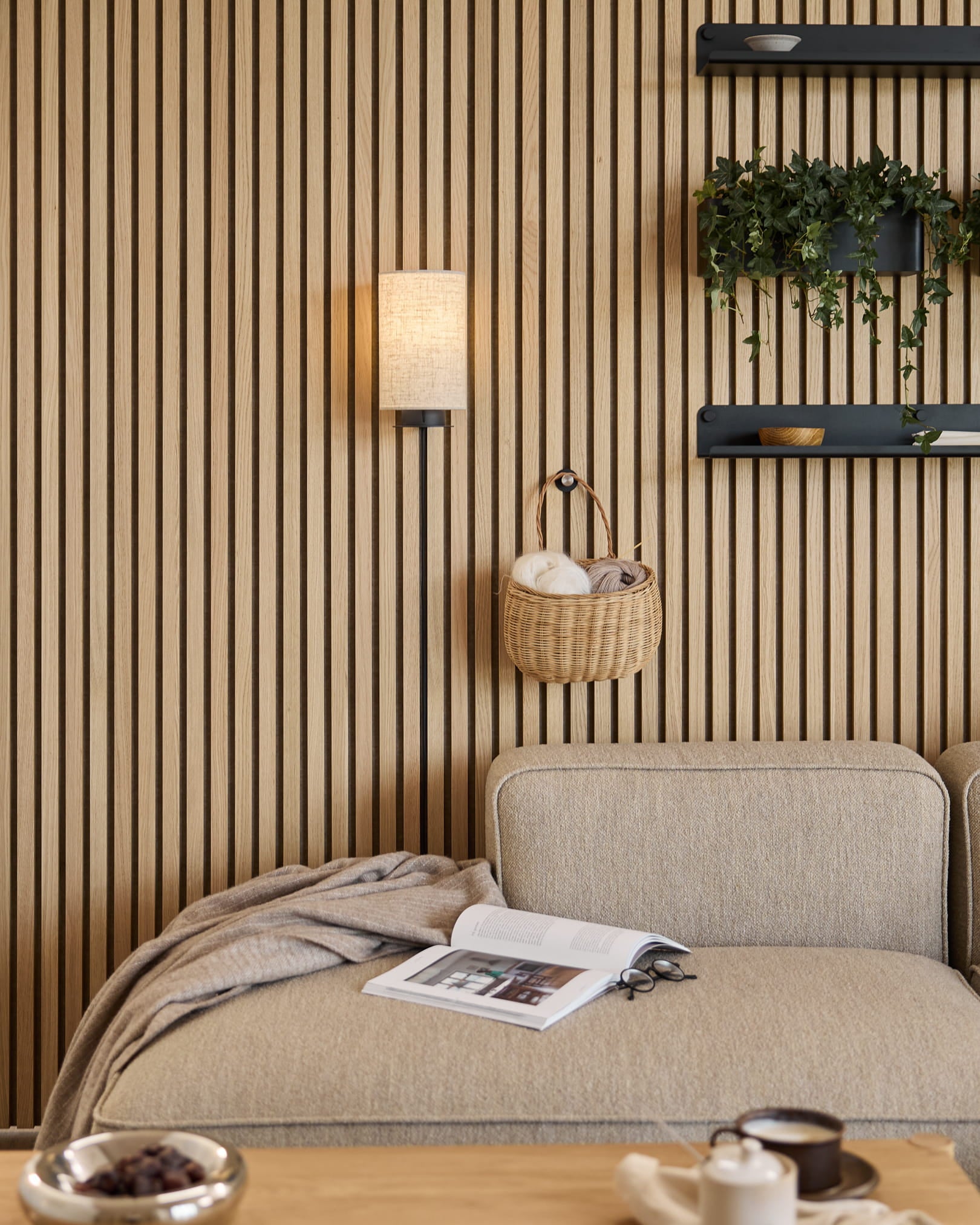 Sofá em Beige com manta e livro, ao lado de uma parede acústica ripada WoodUpp em Akupanel, prateleiras com plantas e peças decorativas, e um candeeiro de parede Feya em Black/Beige.