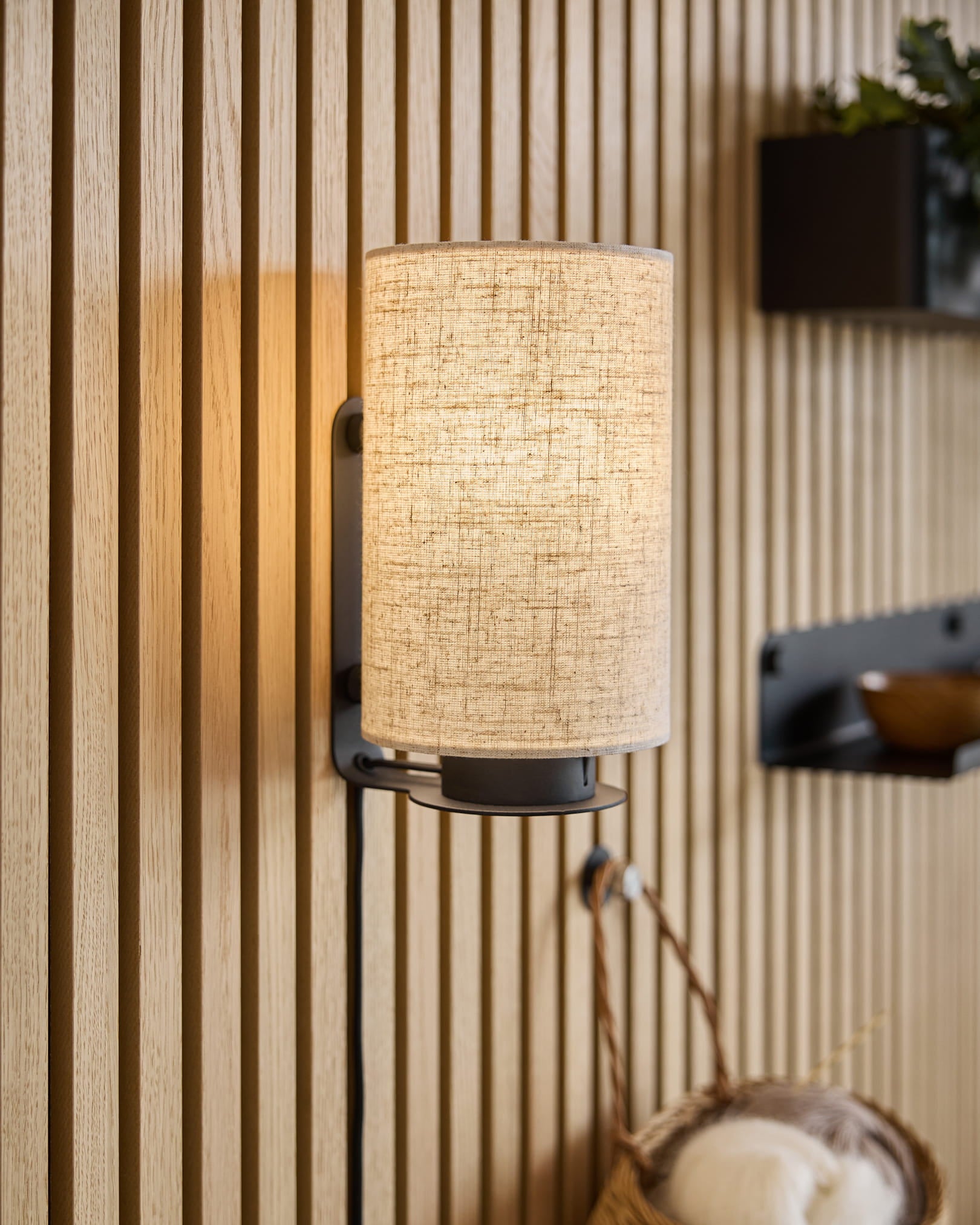 Uma arandela de parede moderna com um abajur de tecido projeta luz quente sobre painéis de madeira acústica Akupanel texturizada, complementada por prateleiras minimalistas e uma cesta de tecido.