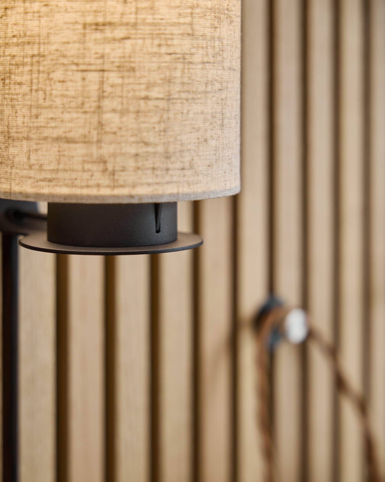 Candeeiro de parede Feya / Black/Beige da WoodUpp com abajur em linho e algodão em beige, base metálica em black e cabo visível, montado num painel acústico ripado de inspiração nórdica.