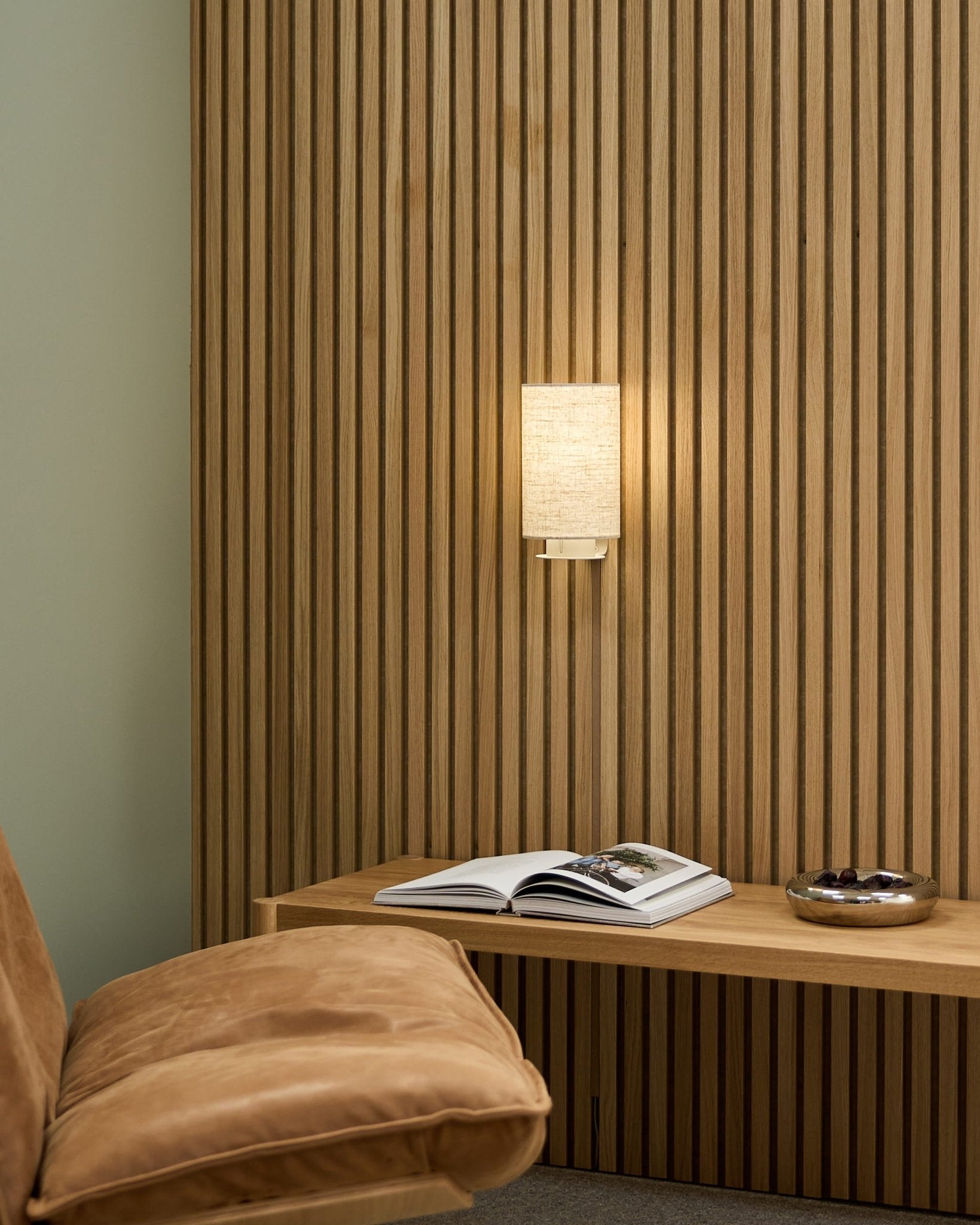 Espaço nórdico minimalista com uma cadeira em tom camel, parede acústica ripada WoodUpp Akupanel, candeeiro de parede Feya em Beige/Beige, um livro aberto sobre um banco e uma taça.