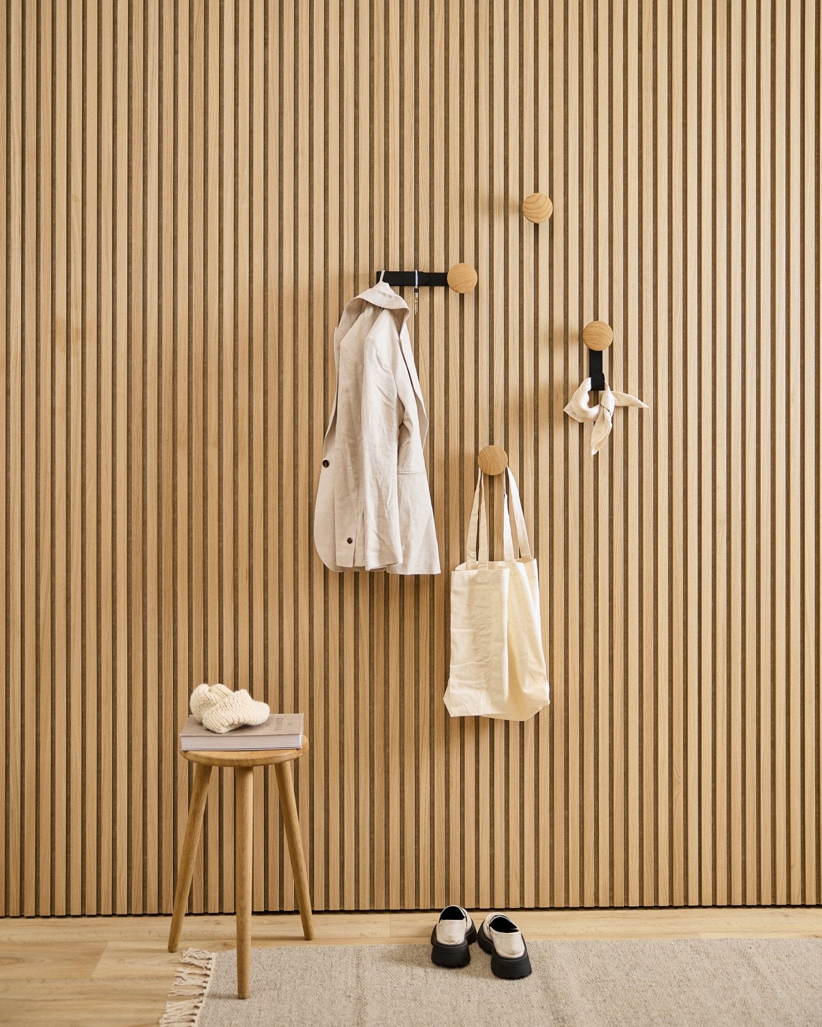 Entrada minimalista com o cabide WoodUpp Linn Vertical em natural oak, com um casaco em beige, saco tote, cachecol, banco com chinelos e sapatos em black e branco.
