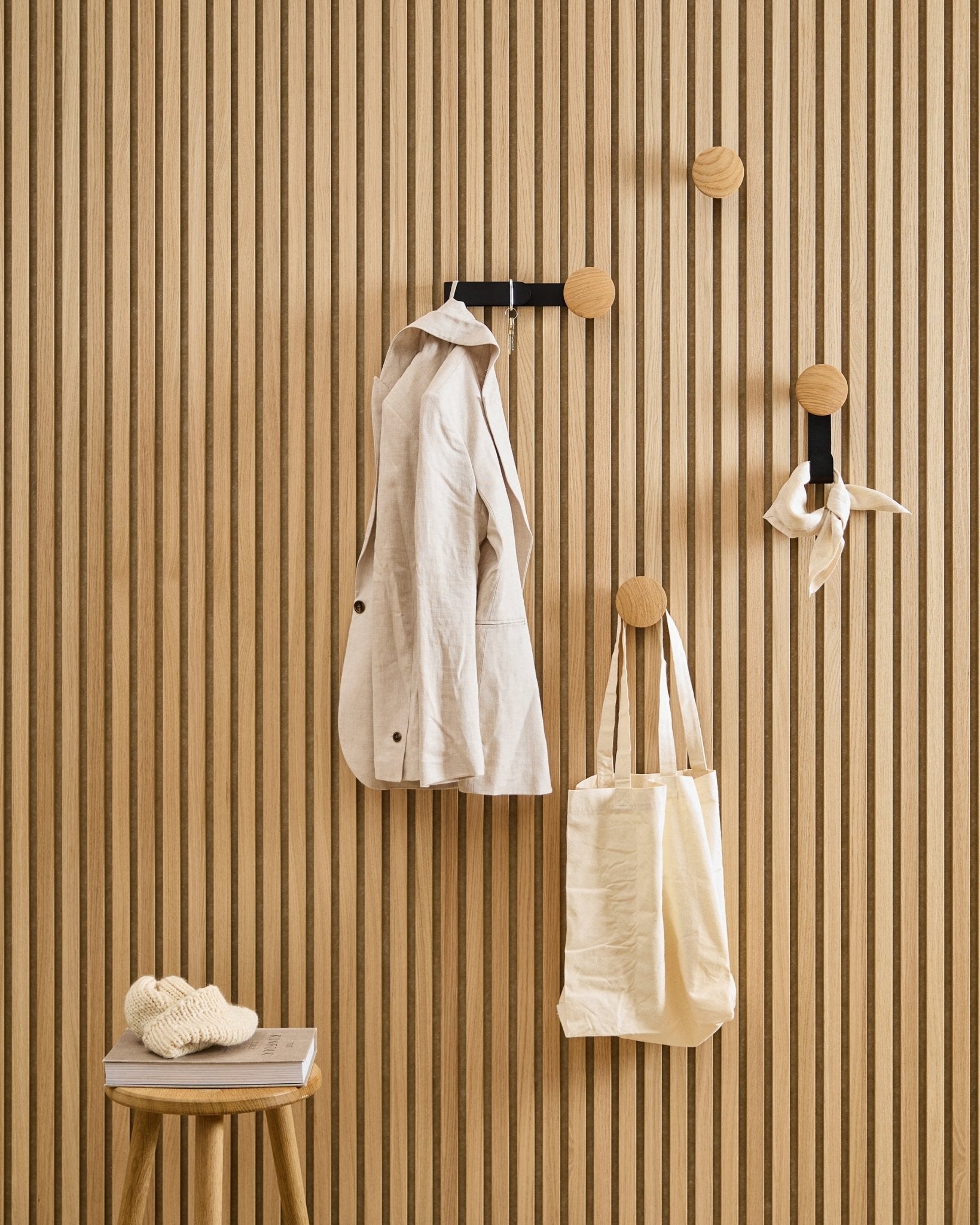 Um casaco em beige, um saco tote e um cachecol estão pendurados no Hanger Linn Horizontal da WoodUpp em Natural Oak; um banco ao lado suporta um livro e uma peça de malha.