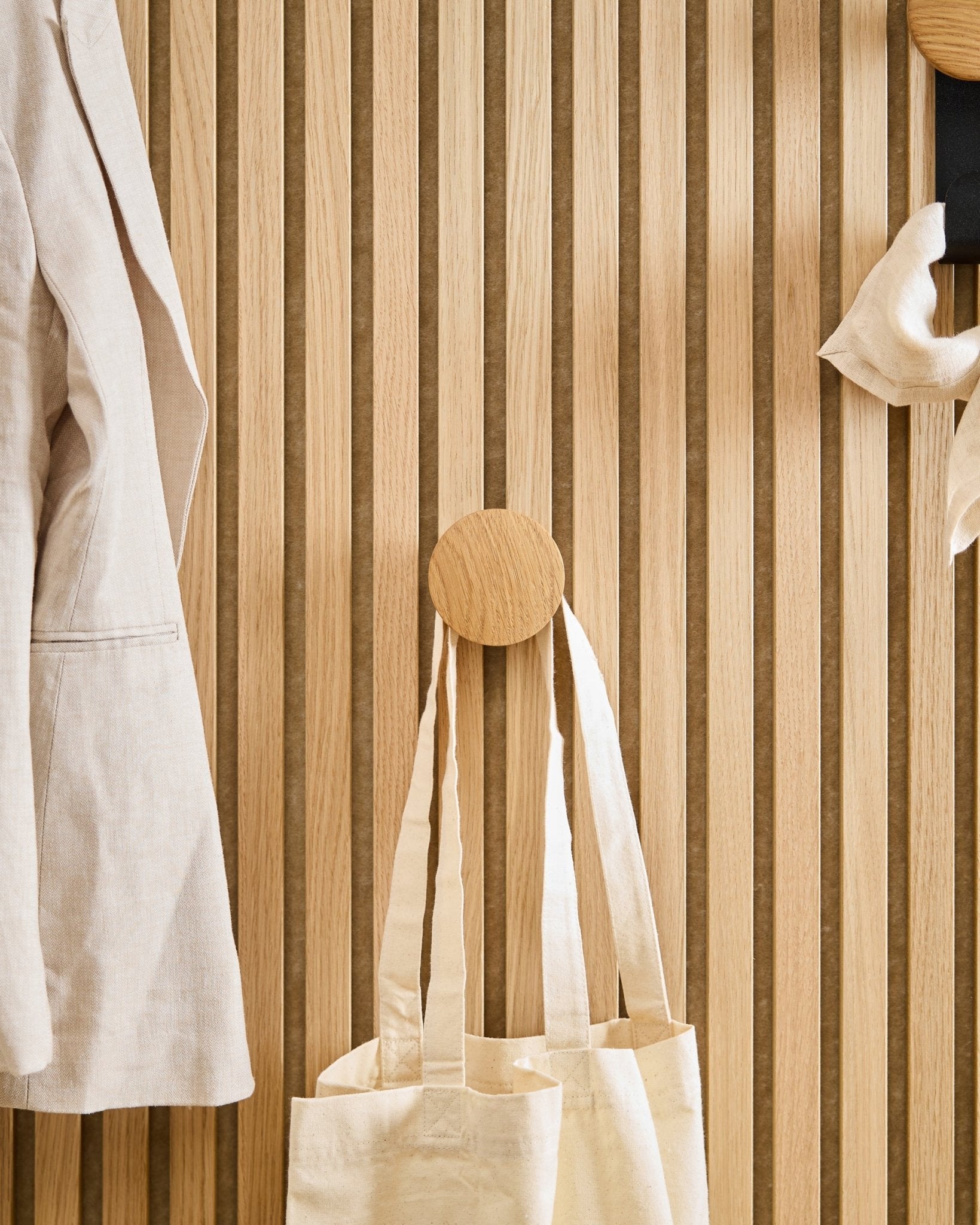 Um casaco beige e um saco tote estão pendurados no Linn I Single Hanger em Natural Oak da WoodUpp, montado num painel acústico de ripas de madeira vertical Akupanel.