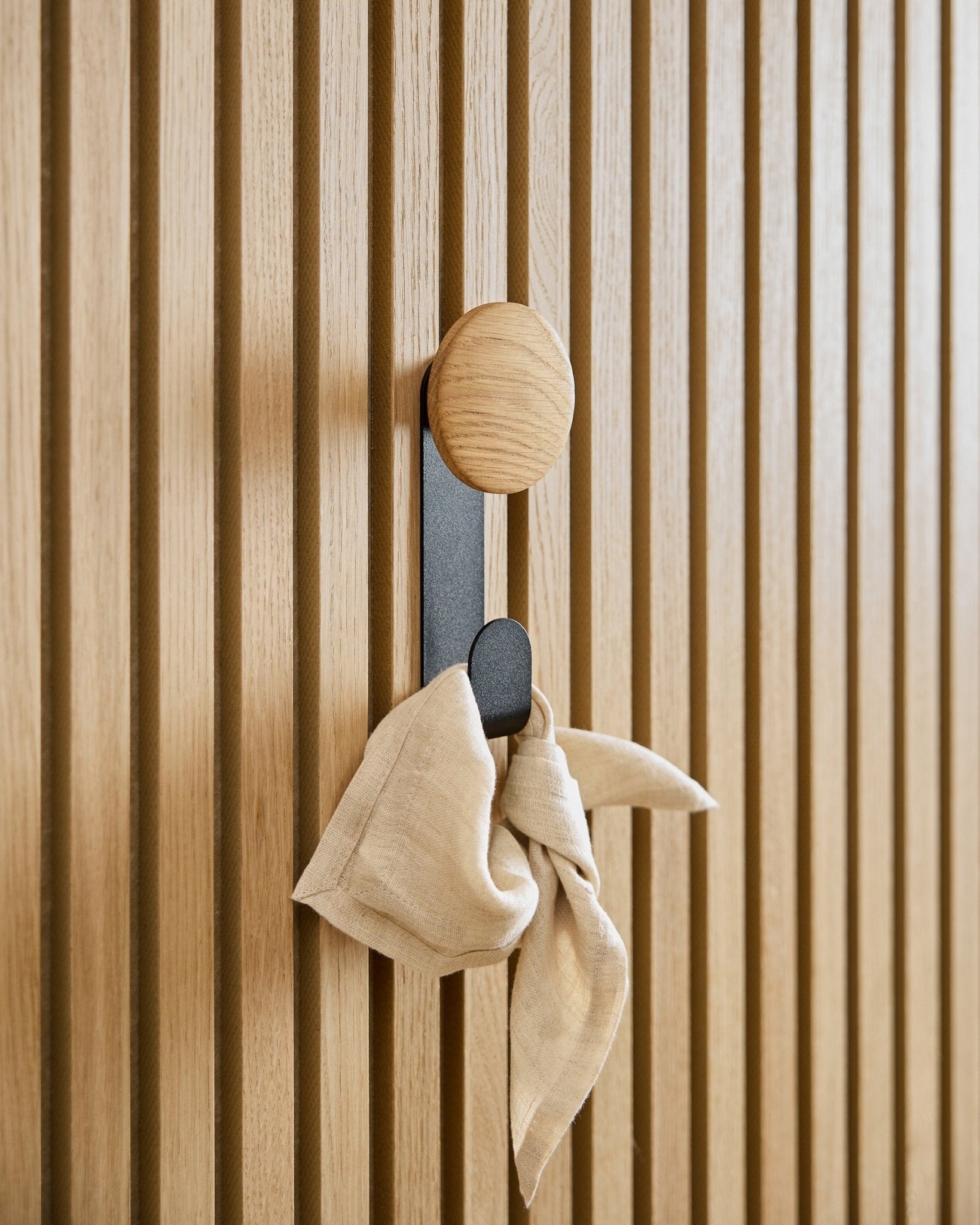 Um pano em beige está pendurado no cabide Linn Vertical em Natural Oak da WoodUpp, montado num painel acústico ripado de parede em vertical Akupanel.