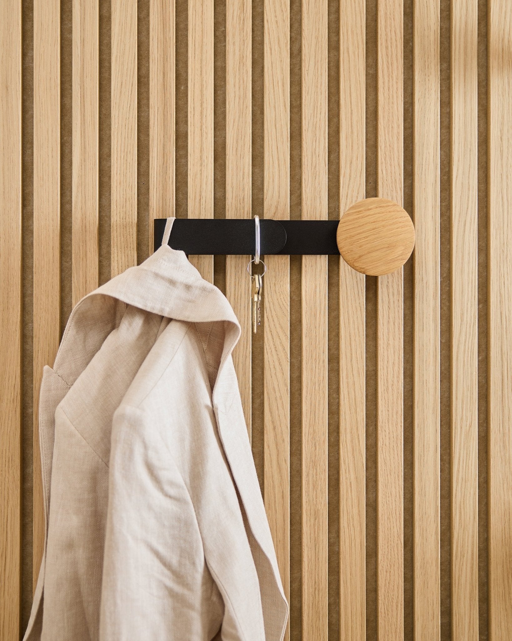 Um blazer em beige e um molho de chaves estão pendurados no Hanger Linn Horizontal da WoodUpp em natural oak, instalado num painel acústico de madeira natural oak Akupanel.