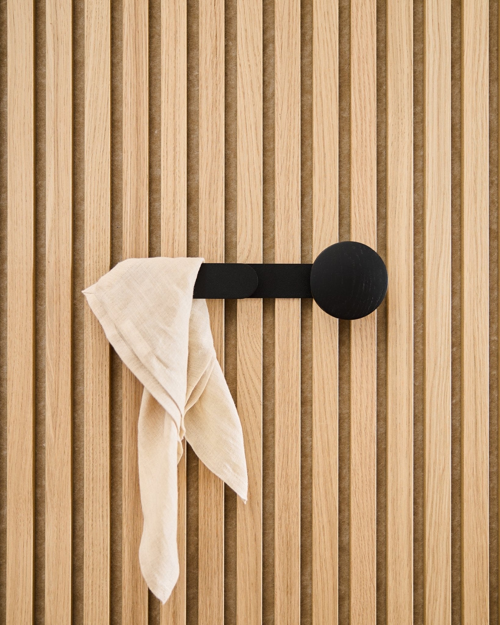 Um tecido beige cai sobre o cabide WoodUpp Linn Horizontal em Black Oak, fixado num painel acústico ripado vertical.