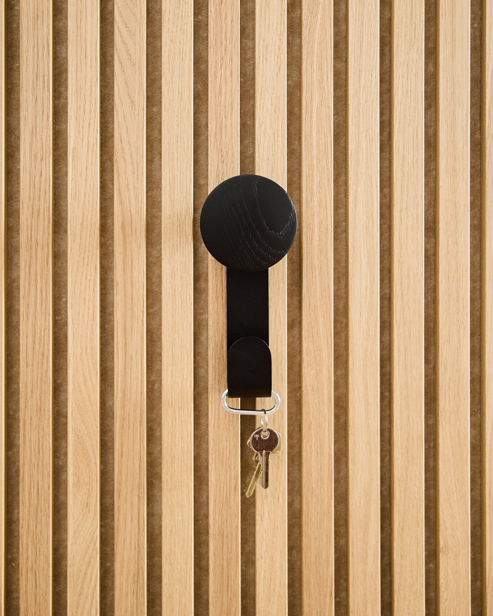 Um cabide Linn Vertical em Black Oak da WoodUpp segura um porta-chaves com duas chaves e uma etiqueta redonda, montado num painel acústico de madeira Akupanel com textura.