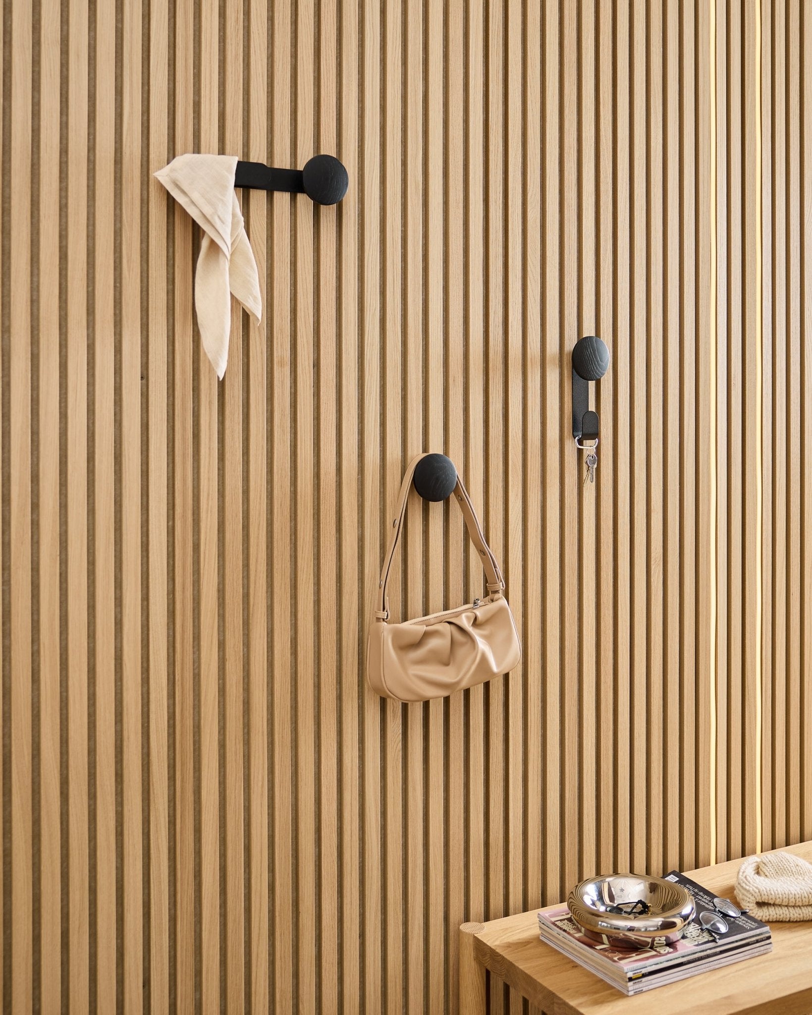 O Single Hanger Linn da WoodUpp em Black Oak segura um cachecol, uma mala beige e chaves num painel acústico ripado de madeira Akupanel black; banco com livros e prato metálico por perto.