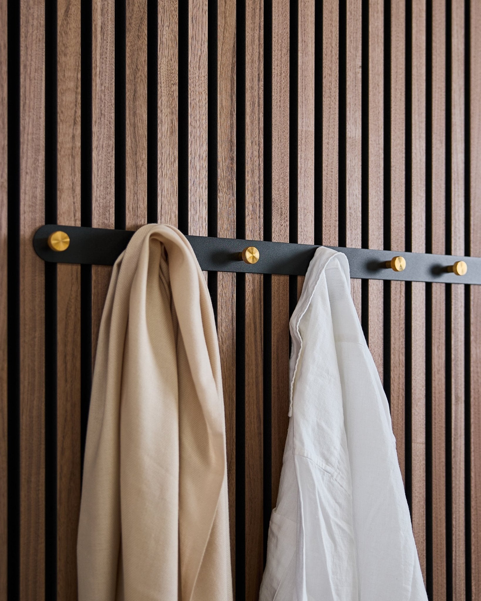 O bengaleiro Coat Hook Rack da WoodUpp Norr em black/brass (X7), com um cachecol beige e uma camisa branca, está pendurado num painel acústico de madeira com ripas de estilo nórdico.