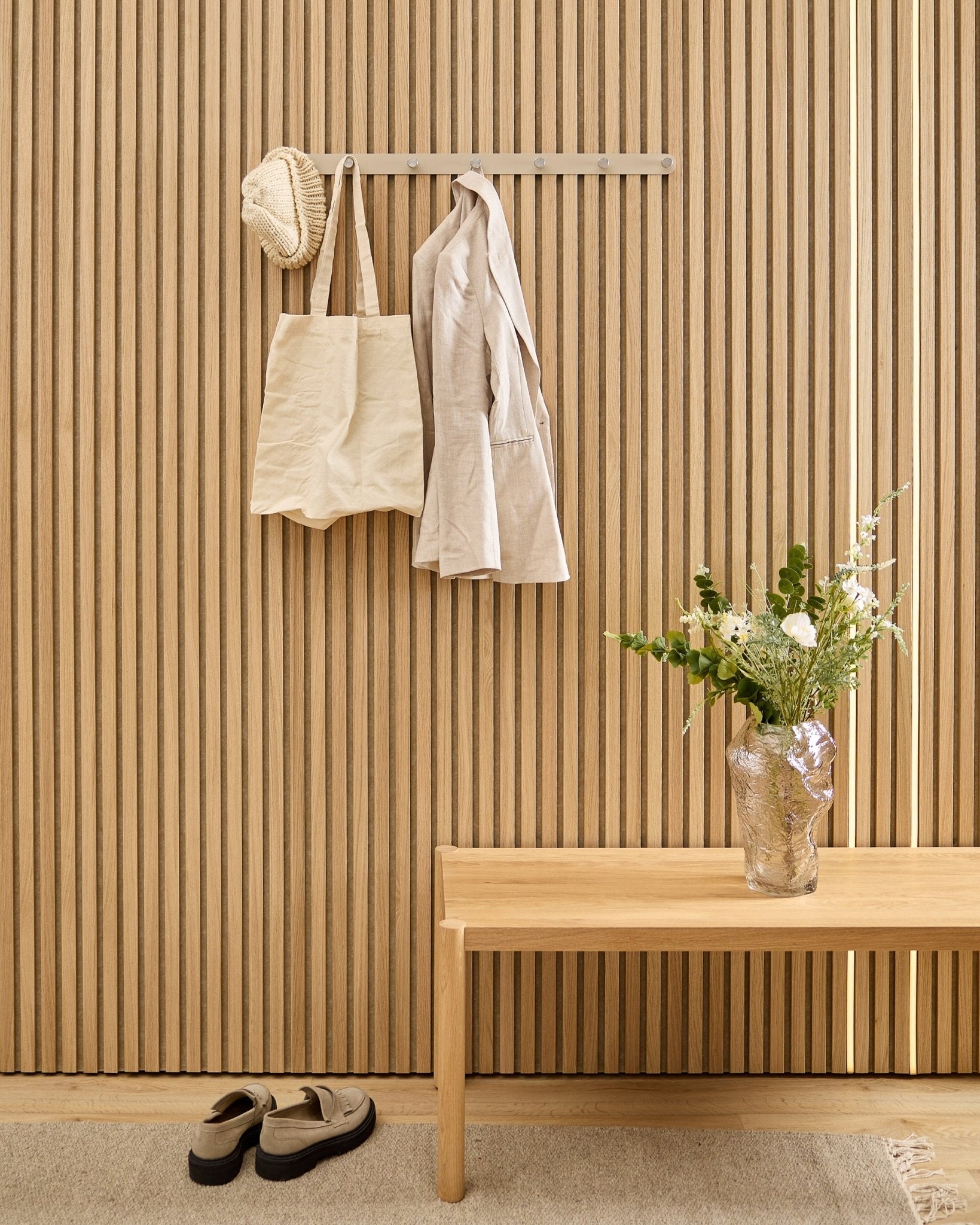 Um saco beige, um chapéu e uma camisa estão pendurados no cabideiro com ganchos WoodUpp Norr, por cima dos sapatos, com um banco e uma jarra ao lado do painel acústico ripado às riscas.