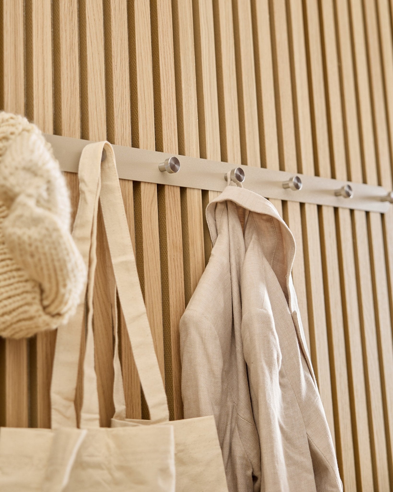 Um saco tote beige, um gorro de malha e uma camisa de linho estão pendurados no cabideiro com ganchos WoodUpp Norr (Beige/Aço inoxidável Steel, X7) montado num painel ripado Akupanel em madeira clara.