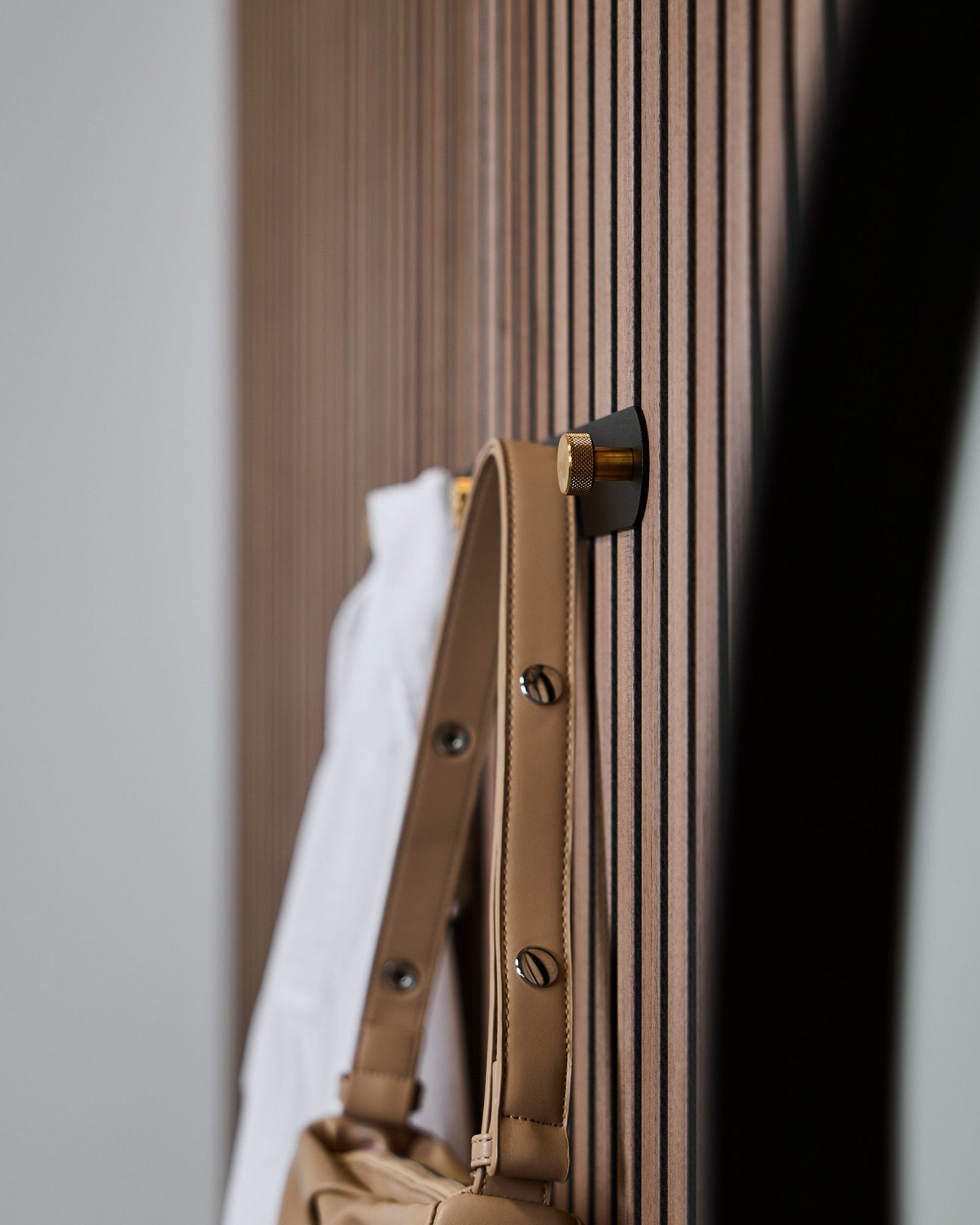 Uma mala castanha e uma camisa branca estão penduradas num cabide de parede WoodUpp Norr (Black/Brass, X5), montado num painel acústico ripado vertical Akupanel.