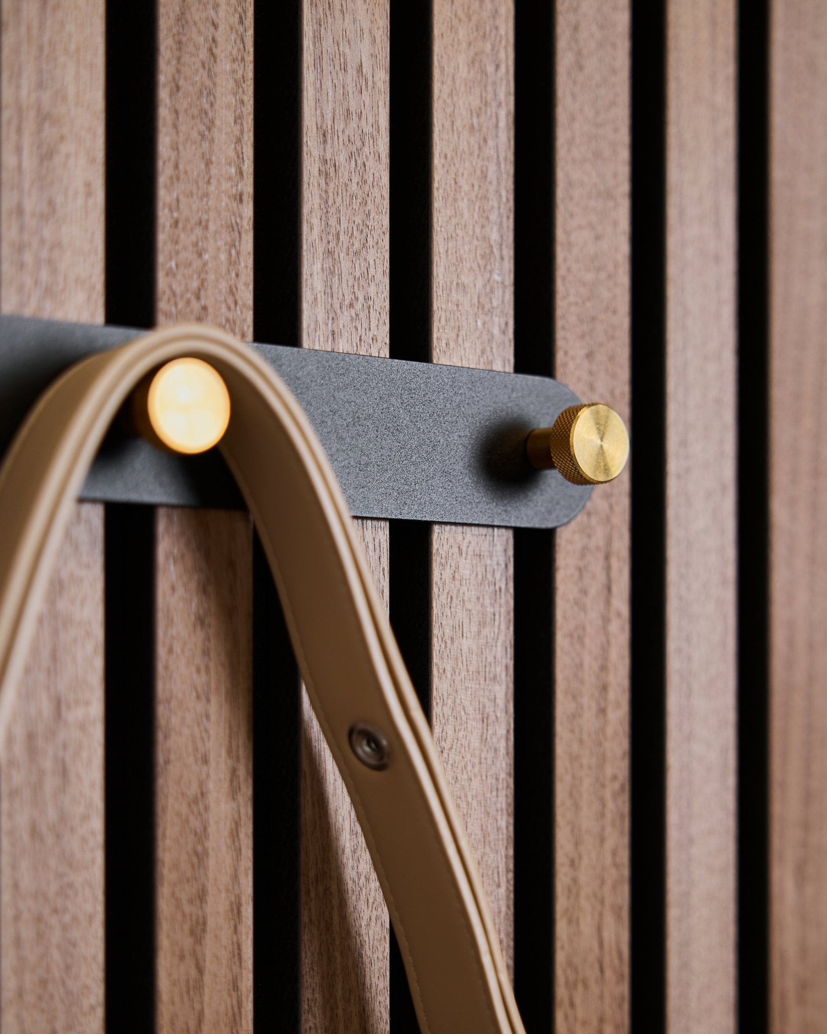 Cinco ganchos black/brass do cabide de parede Norr da WoodUpp, montado num painel acústico ripado em madeira, cada um com uma alça beige pendurada.