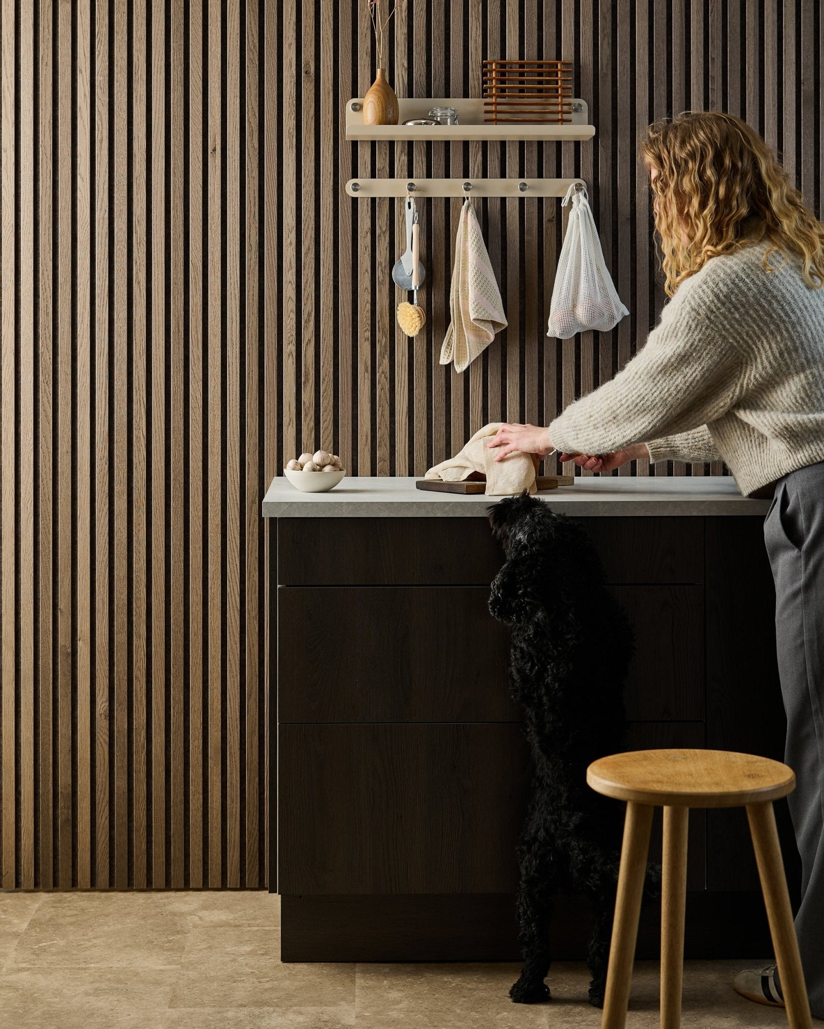 Um cão black está apoiado nas patas traseiras, a tentar alcançar comida numa bancada de cozinha, com painéis acústicos de madeira Nordic e prateleiras ao fundo.
