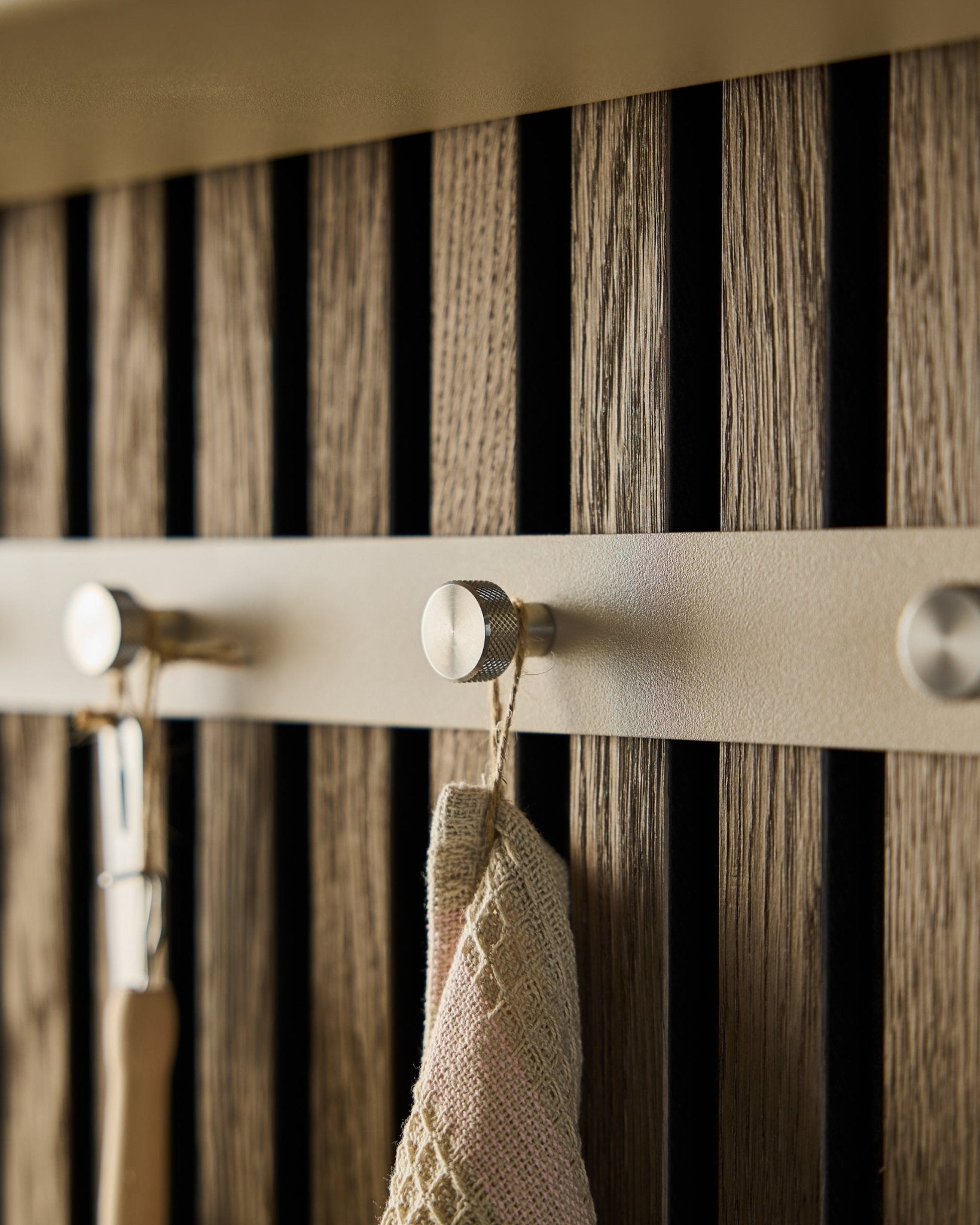 Plano aproximado do cabideiro de parede Norr da WoodUpp (Beige/Aço inoxidável Steel, X5) montado num painel de ripas de madeira Painel acústico, com uma toalha e uma ferramenta com cabo penduradas.