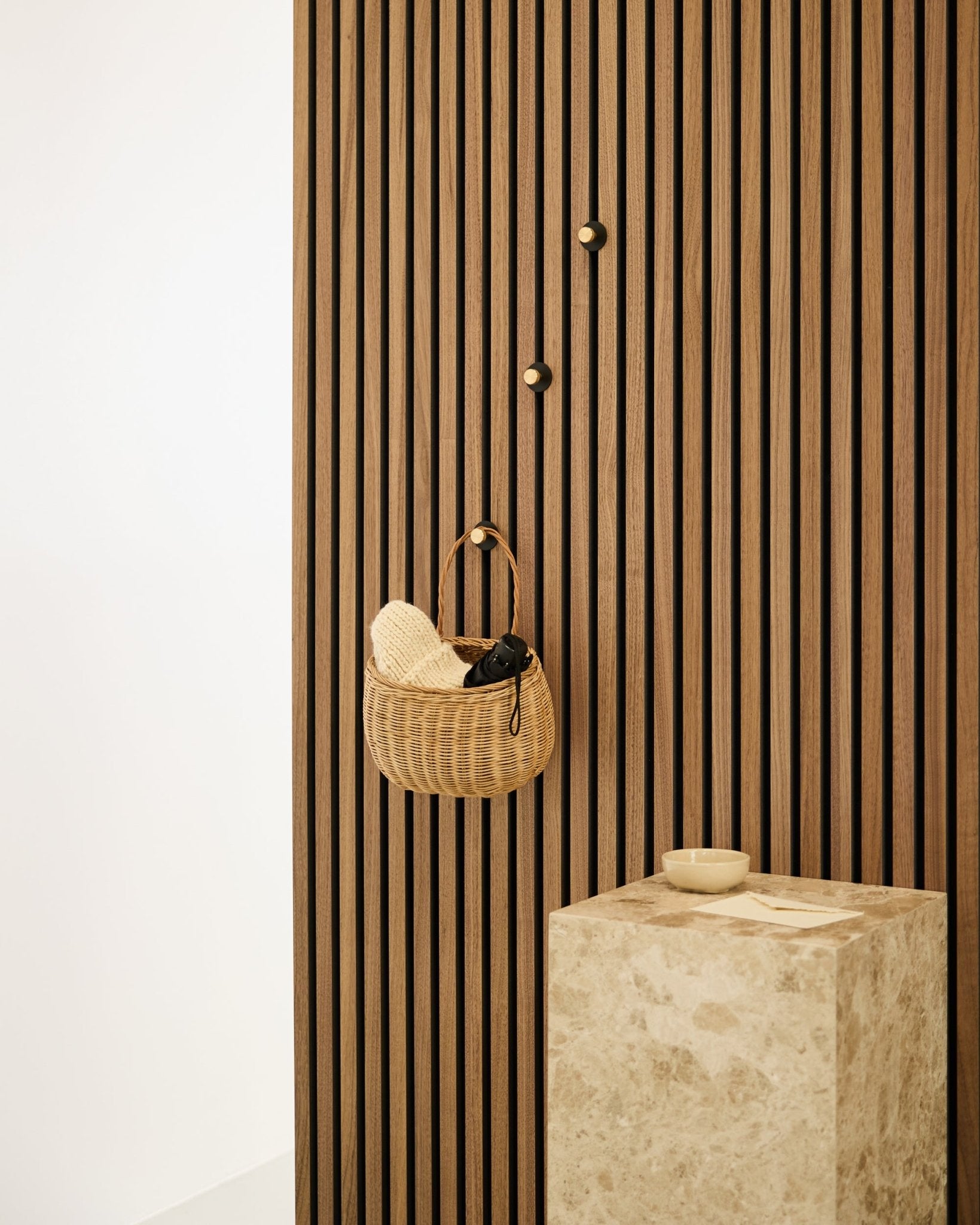 Gancho de parede Norr da WoodUpp montado numa parede acústica de ripas nórdica Akupanel, ao lado de um pedestal de pedra beige com uma taça small, num interior minimalista.