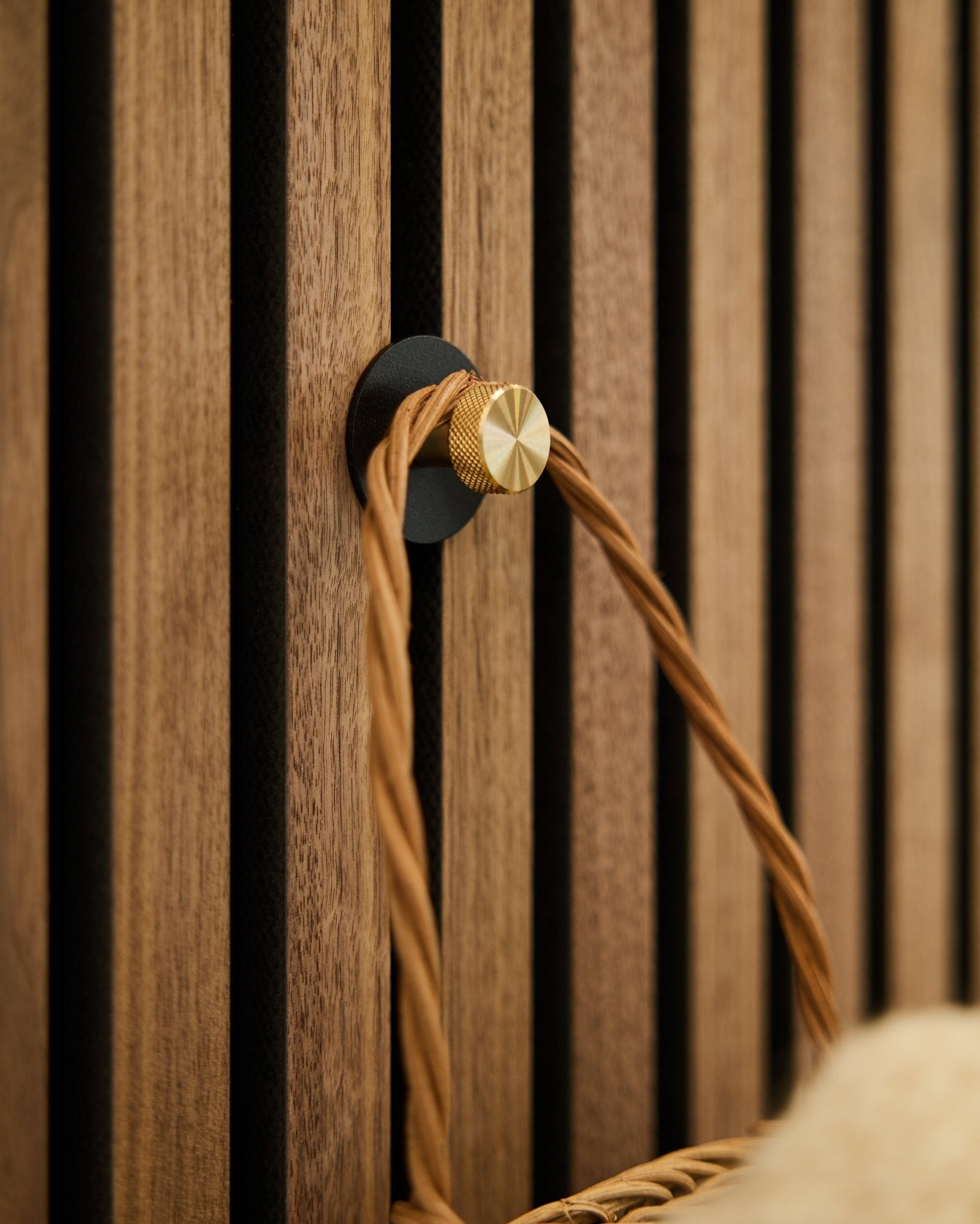 Gancho de parede WoodUpp Norr em black e brass, montado numa base de madeira Akupanel Painel acústico, segura uma pega entrançada em tom bege.