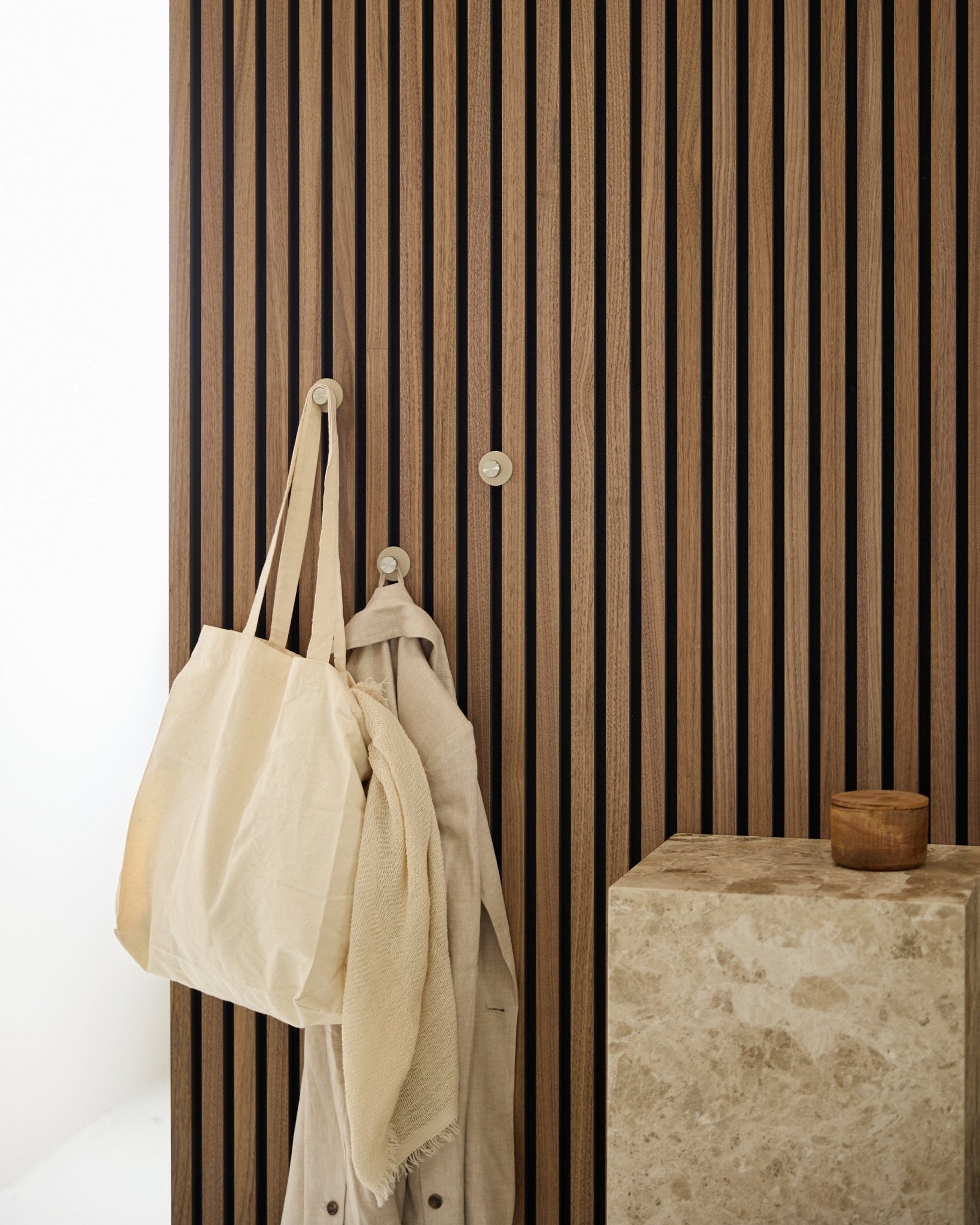 Um saco tote beige e um tecido leve estão pendurados num cabide WoodUpp Norr (Beige/Steel inoxidável) montado num vertical Akupanel Painel acústico, ao lado de uma superfície em pedra.