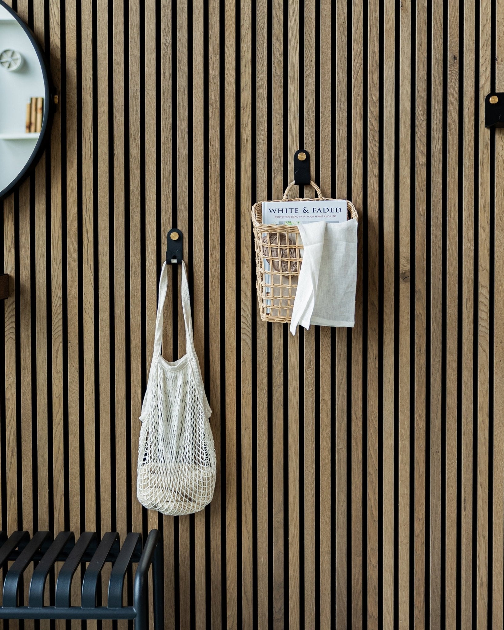 Um saco em rede e um cesto estão pendurados em Single Hangers Norr da WoodUpp num painel acústico ripado de madeira Nordic Akupanel Painel acústico; parte de um espelho é visível à esquerda.
