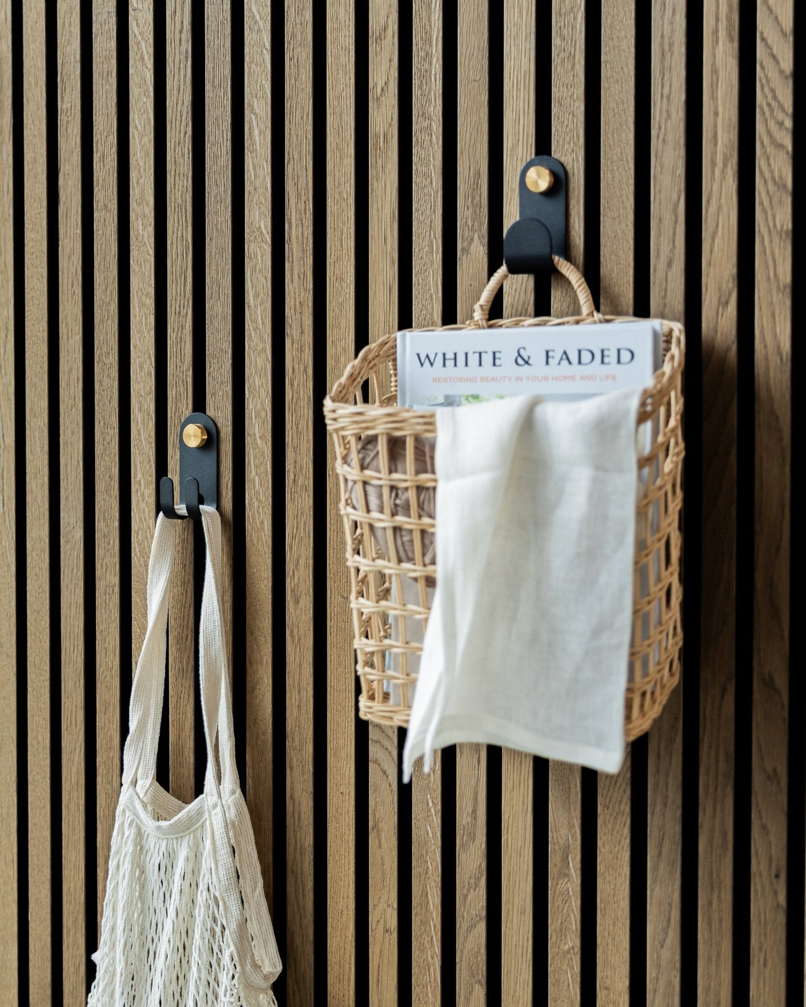 O Norr Single Hanger da WoodUpp em Black, com um cesto entrançado e um saco de rede, está montado num painel acústico ripado nórdico para uma arrumação organizada.