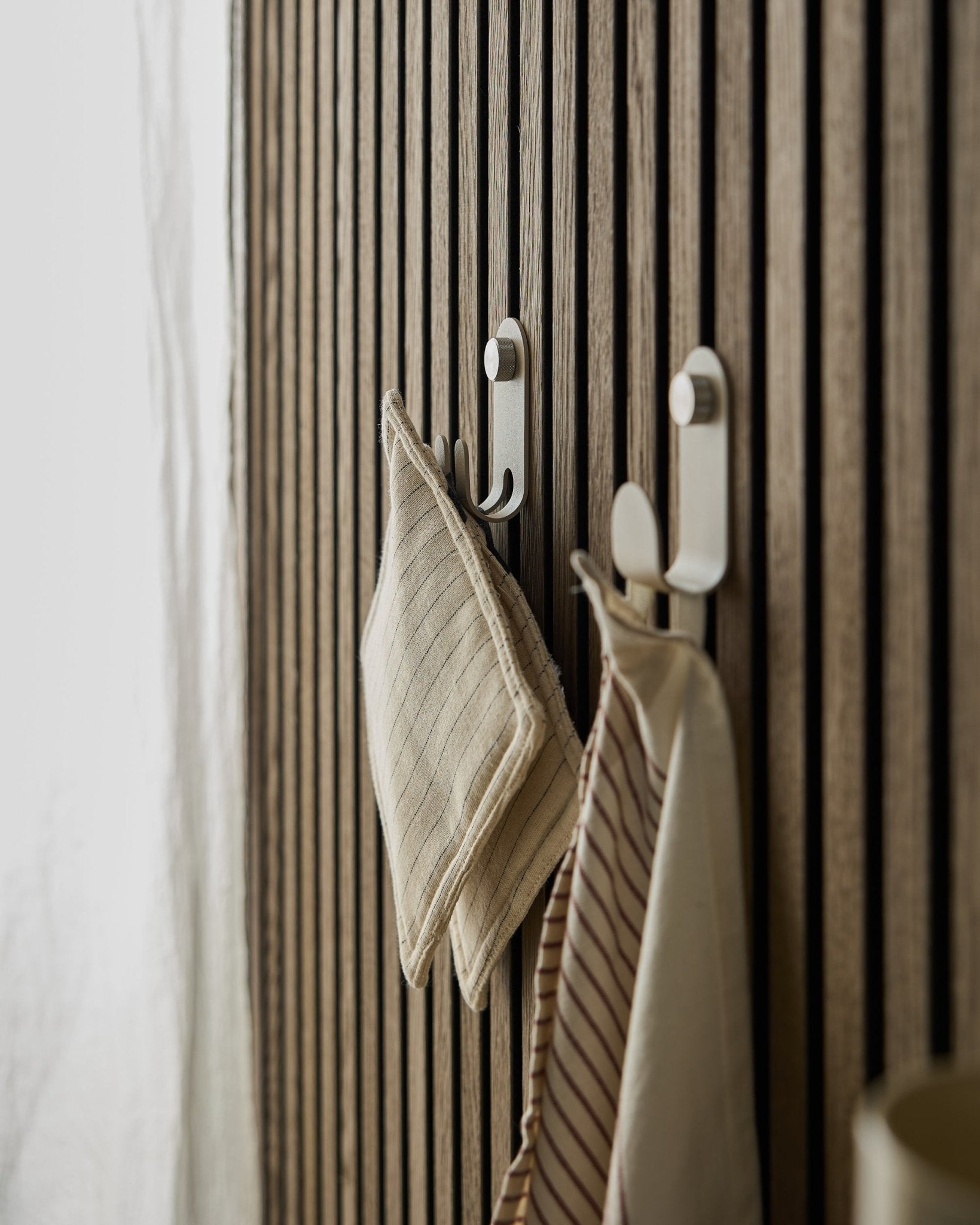 Dois panos de cozinha às riscas estão pendurados no Norr Double Hanger da WoodUpp, montado num painel ripado nórdico beige Painel acústico, iluminado por luz natural suave.