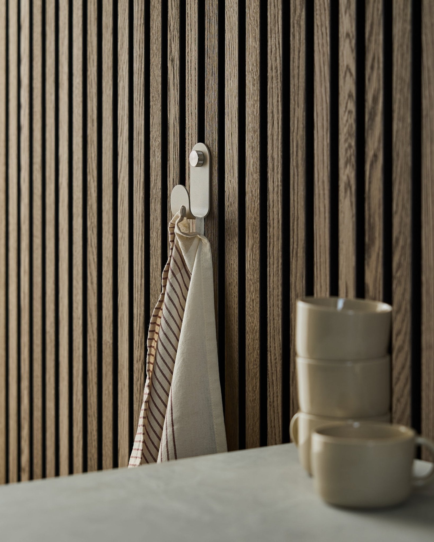 Uma toalha às riscas está pendurada num WoodUpp Norr Single Hanger em beige, montado num painel acústico ripado de estilo nórdico; três canecas beige estão empilhadas à frente.