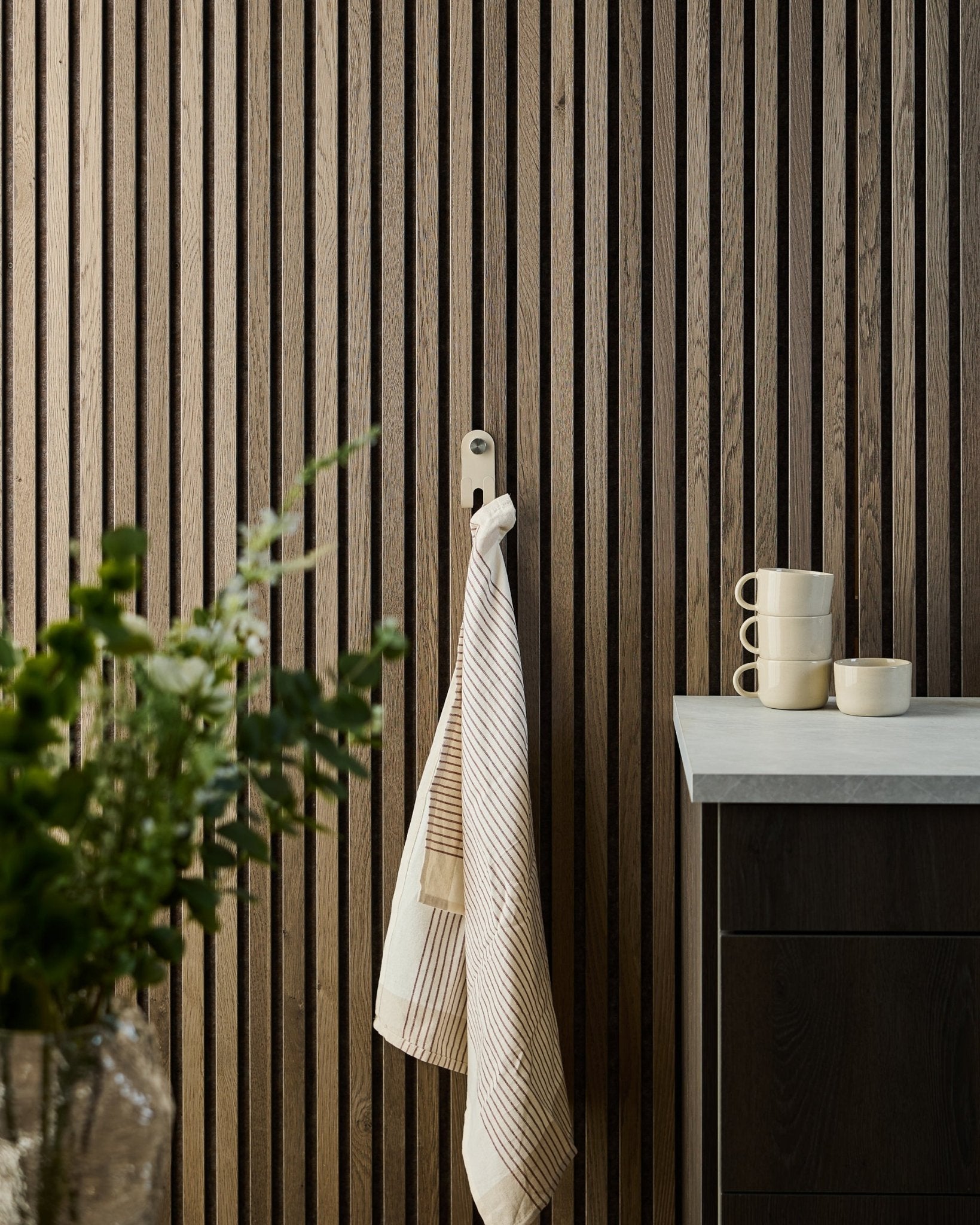 Um pano às riscas está pendurado no WoodUpp Norr Double Hanger em beige, montado num painel ripado acústico nórdico; chávenas, uma taça e uma planta verde estão por baixo.