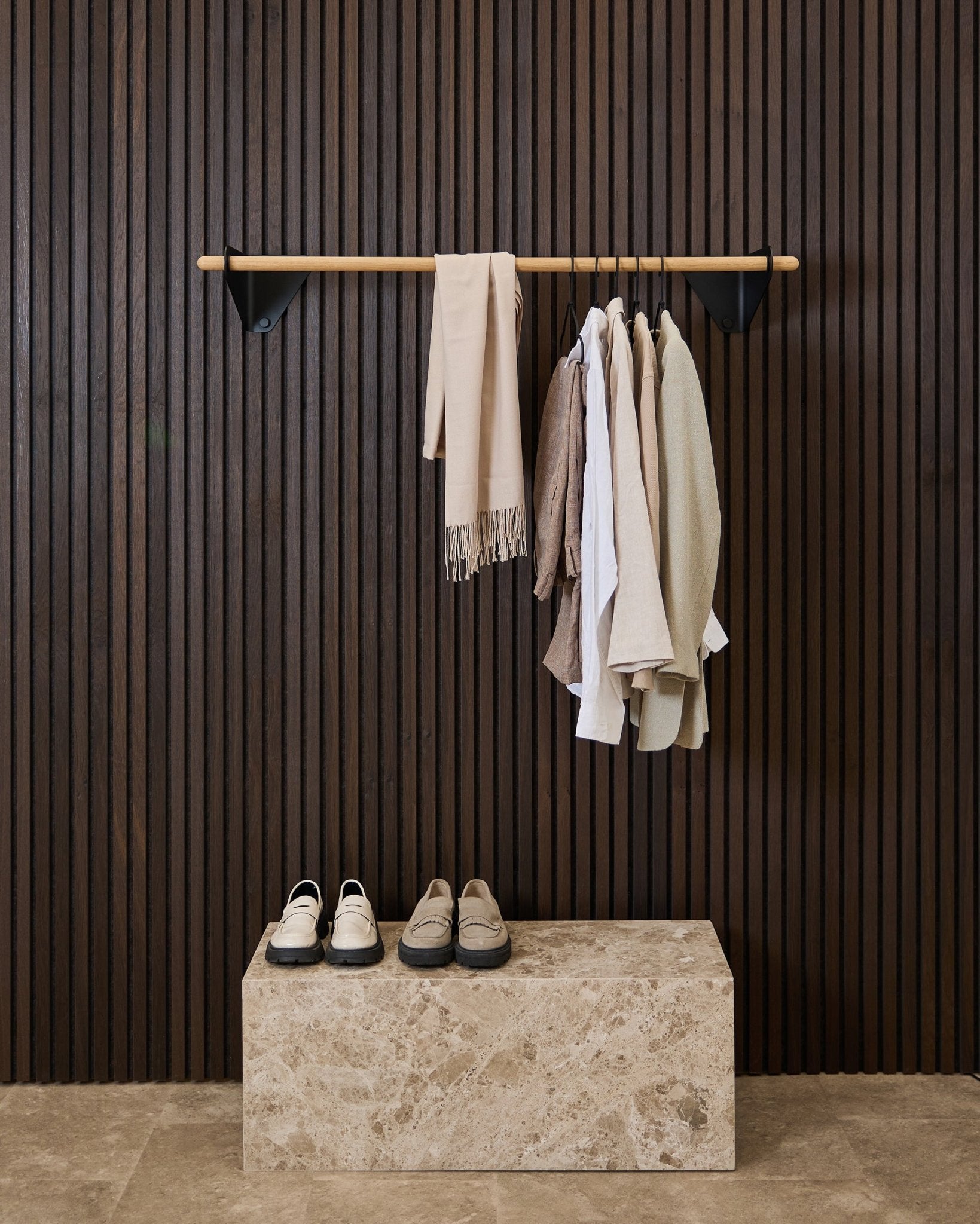 Peças de roupa em tons neutros e um cachecol pendem de um black de 115,9 cm Linn | Coat Rail da WoodUpp, por cima de um banco em mármore, sobre um painel acústico ripado Akupanel escuro.