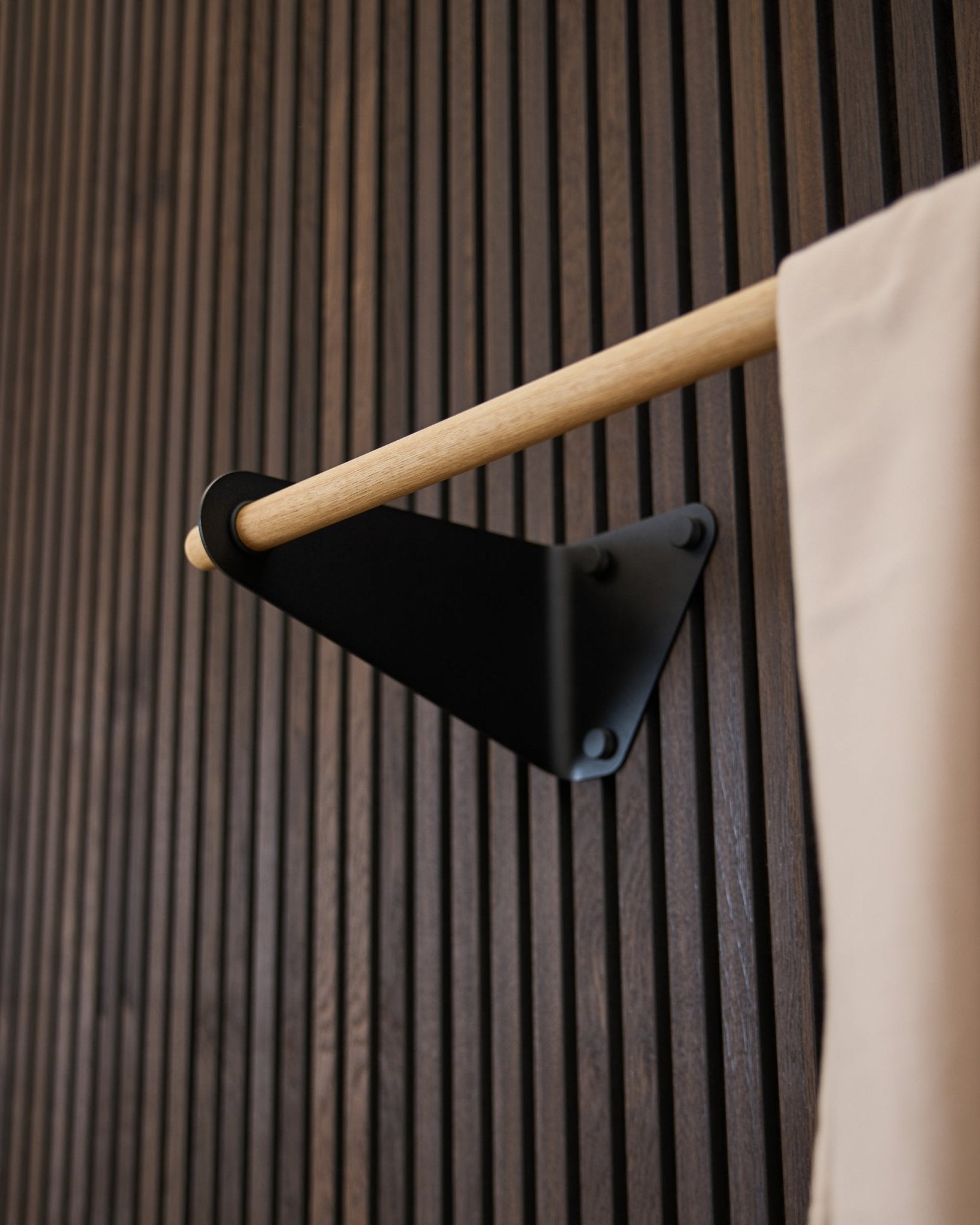 Linn Coat Rail da WoodUpp em black (115,9 cm), montado num painel Painel acústico Akupanel escuro com ranhuras, com detalhe em tecido beige e suporte black.