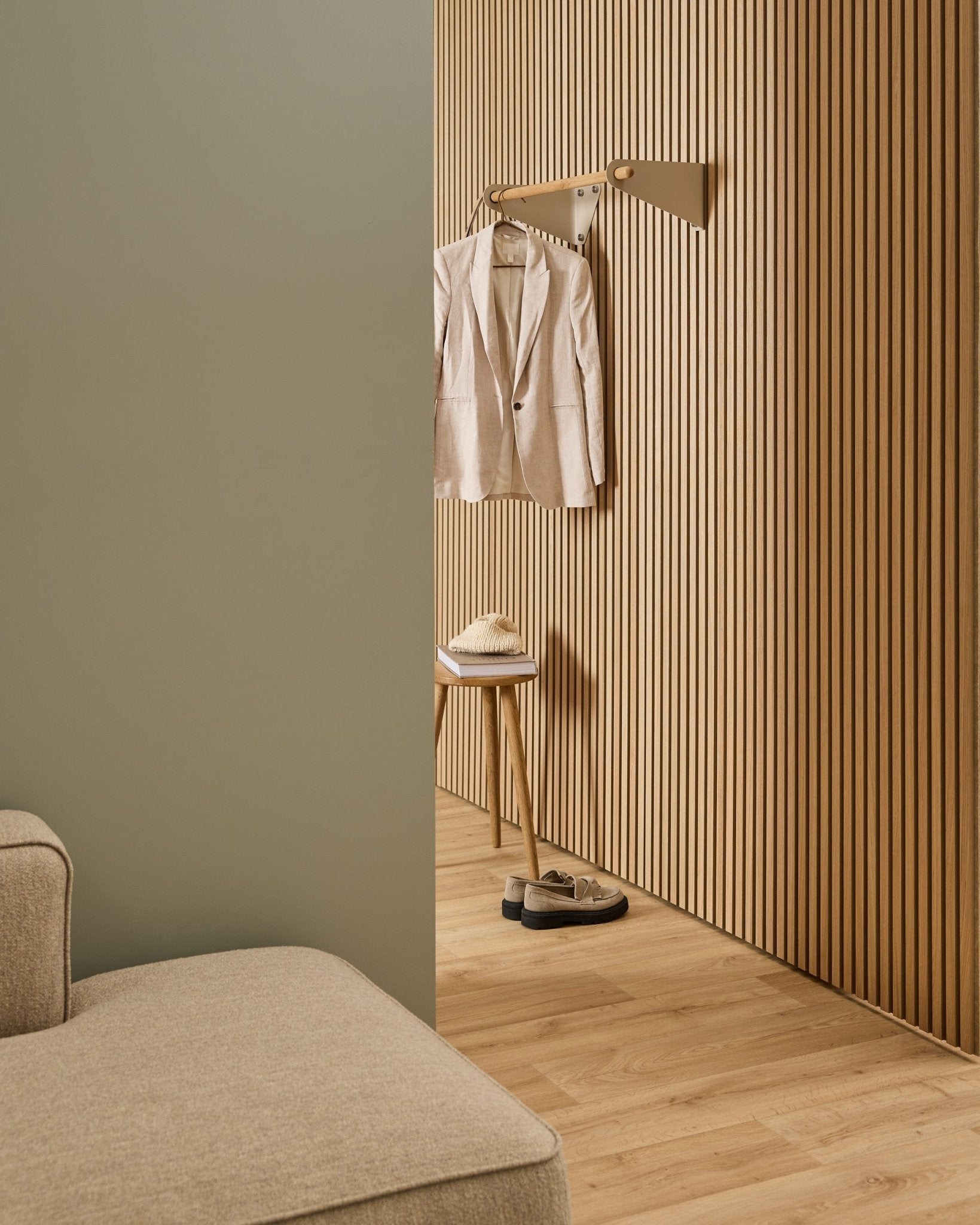 Espaço minimalista com sofá beige, parede acústica ripada Akupanel, cabide WoodUpp Linn Coat Rail em beige (75,9 cm), banco com chapéu e sapatos sobre piso em madeira clara.