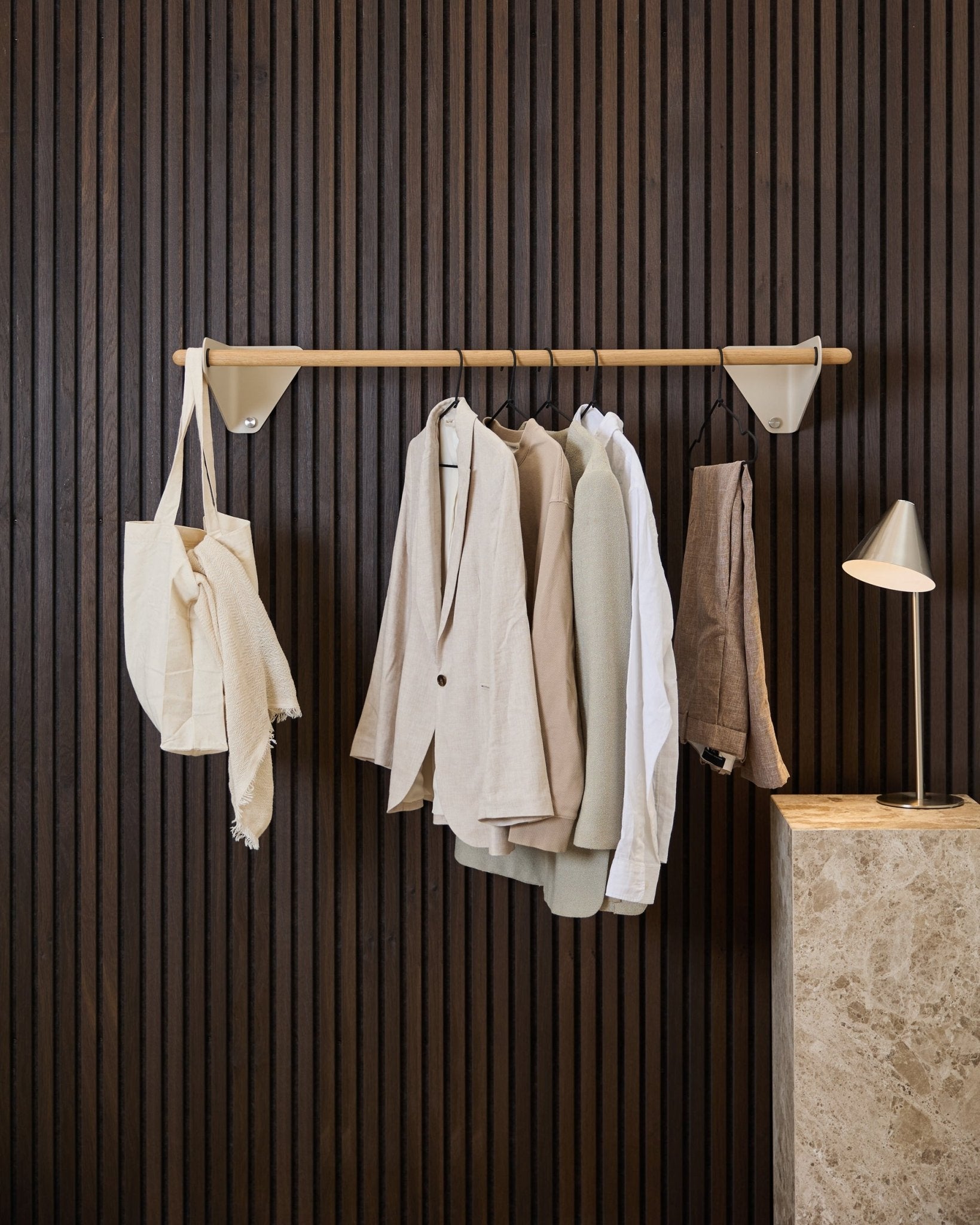 Um cabide WoodUpp Linn em beige (115,9 cm) segura casacos, uma camisa branca e um saco tote em tom creme, em frente a um Akupanel Painel acústico em madeira escura e a um candeeiro de mármore.