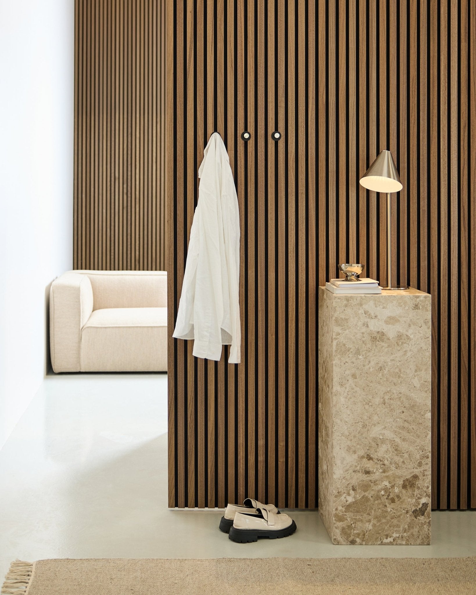 Espaço minimalista com uma parede acústica de ripas de madeira Akupanel de estilo nórdico, camisa branca em ganchos de parede, pedestal em pedra com candeeiro e livros, sofá beige e sapatos brancos no chão.