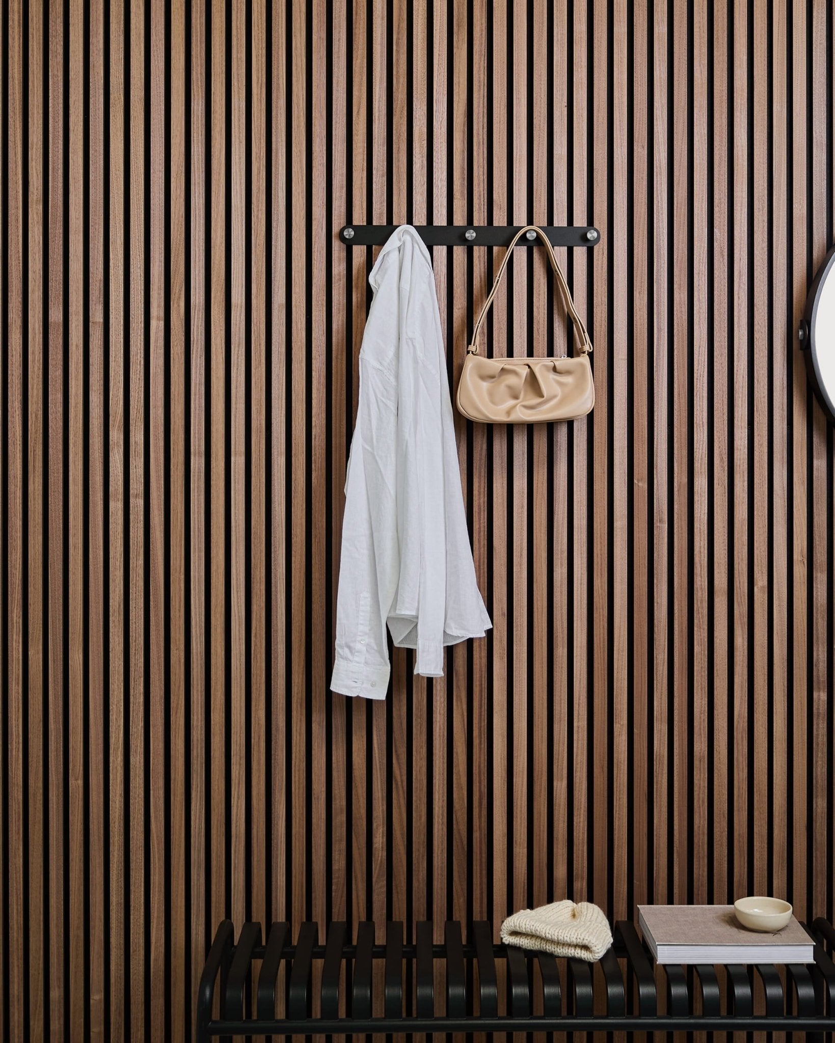 Uma camisa branca e uma mala de mão beige estão penduradas no cabide de parede Norr da WoodUpp, por cima de um banco, contra um painel ripado de madeira Akupanel com peças decorativas.