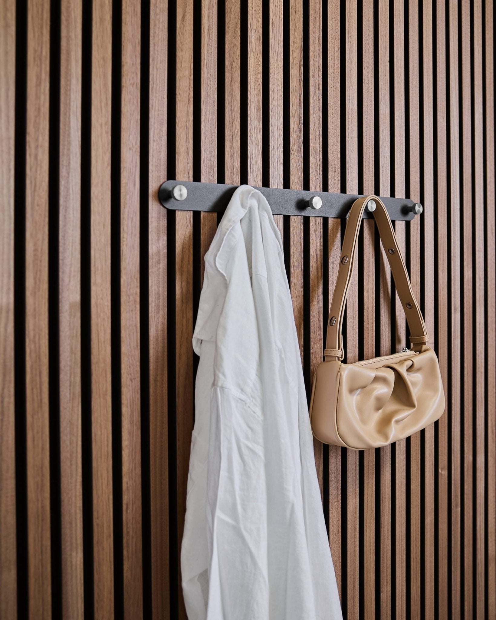Uma camisa branca e uma mala de mão beige estão penduradas nos ganchos de parede Norr X5 Black/Stainless Steel da WoodUpp, fixados a um painel acústico ripado vertical.