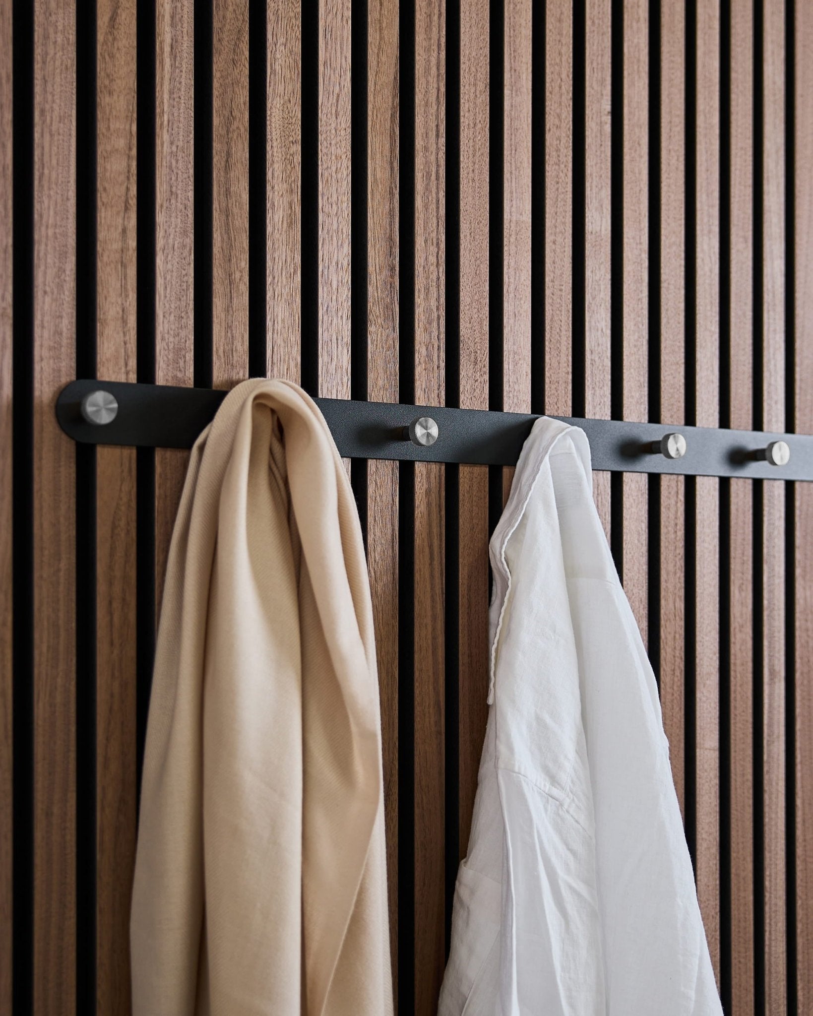 Um lenço beige e uma camisa branca estão pendurados no bengaleiro WoodUpp Norr X7 Black/Stainless Steel, montado num painel acústico ripado de inspiração nórdica.