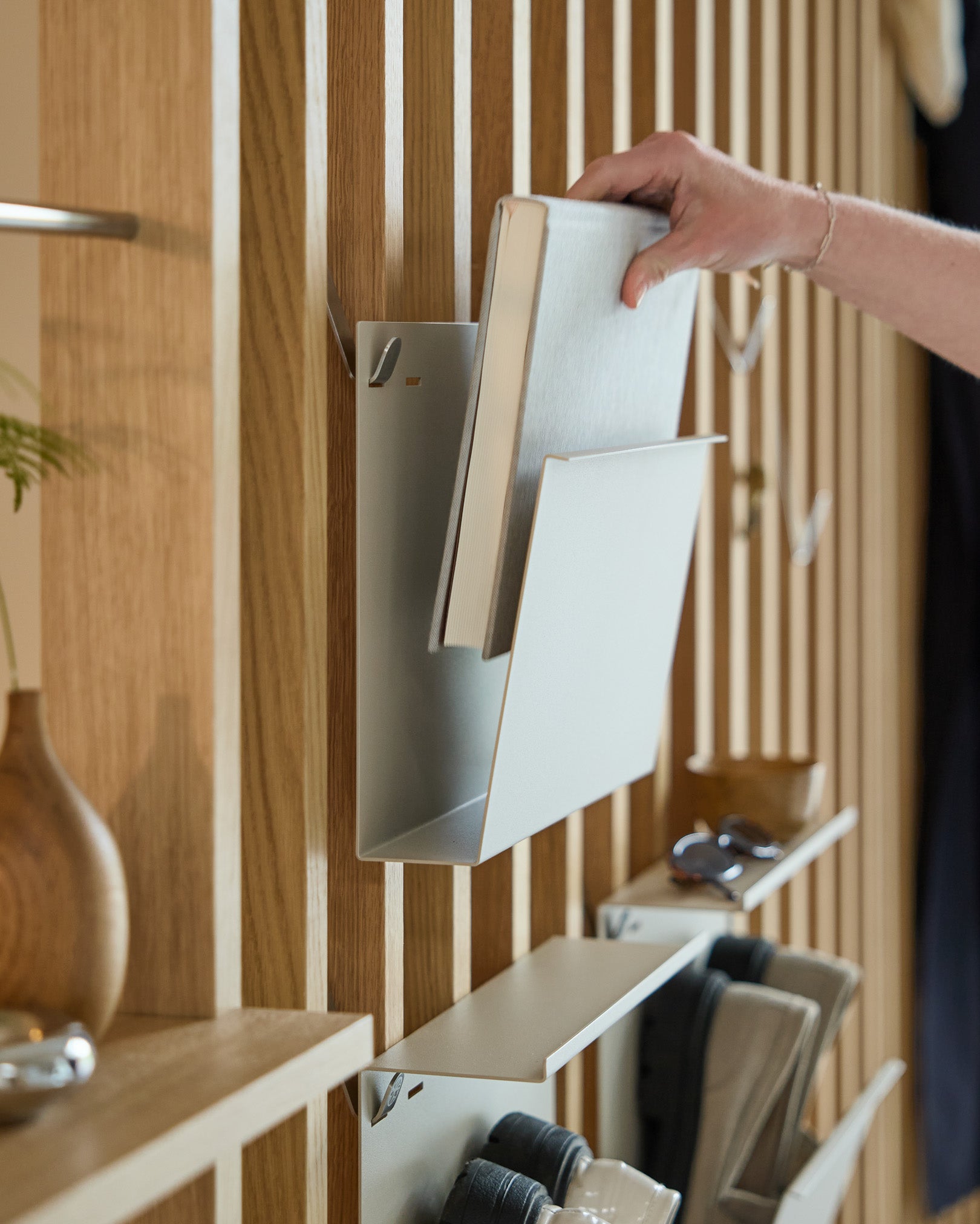 Uma mão coloca Nell | Magazine Storage / Beige da WoodUpp numa prateleira em steel com revestimento a pó, montada num Painel acústico nórdico em madeira, com outros artigos por perto.